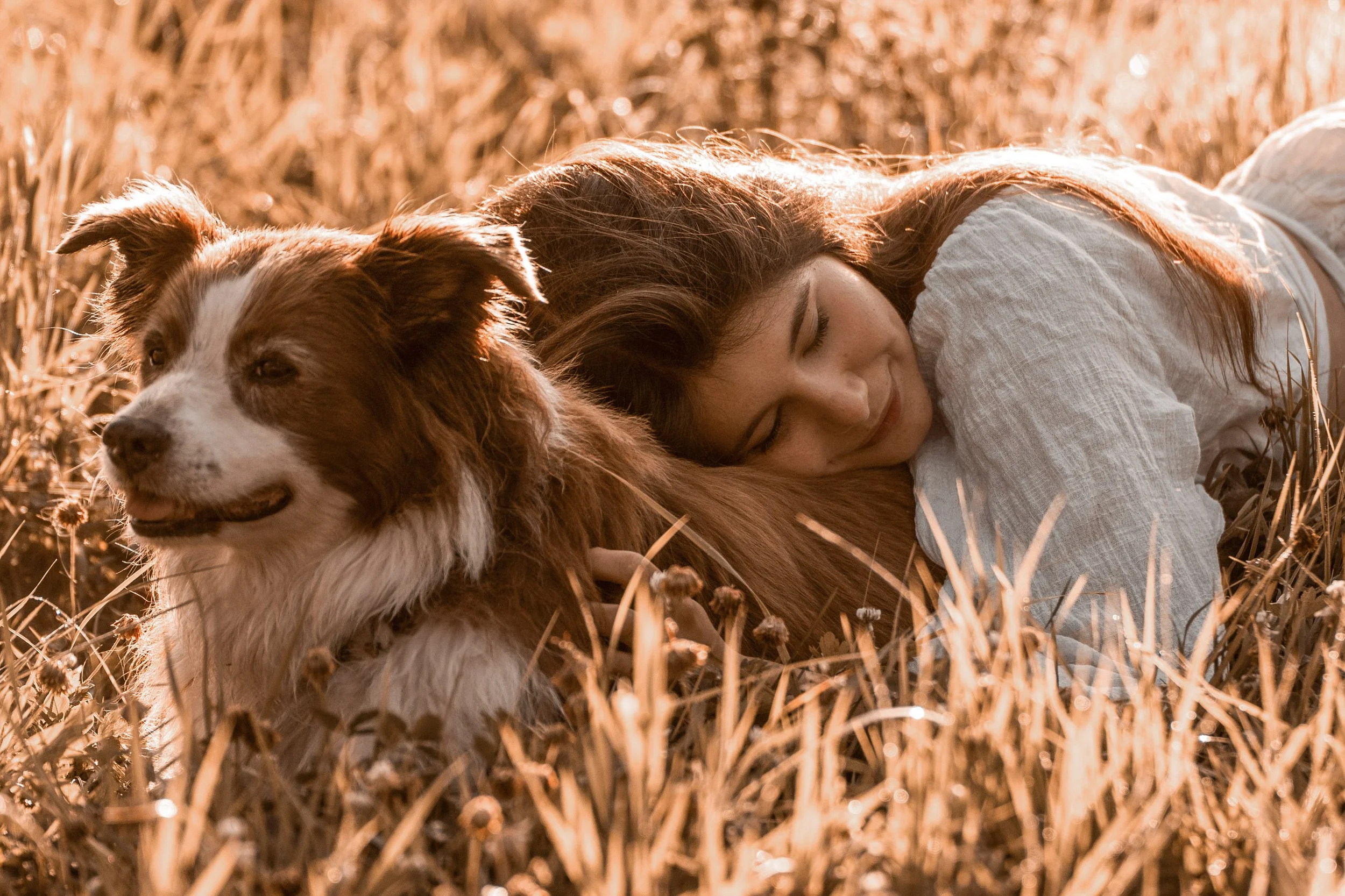 A woman with long red hair lying on her side in a field of dried grass, resting her head on a dog with white and brown fur, both appearing peaceful and relaxed in warm sunlight.