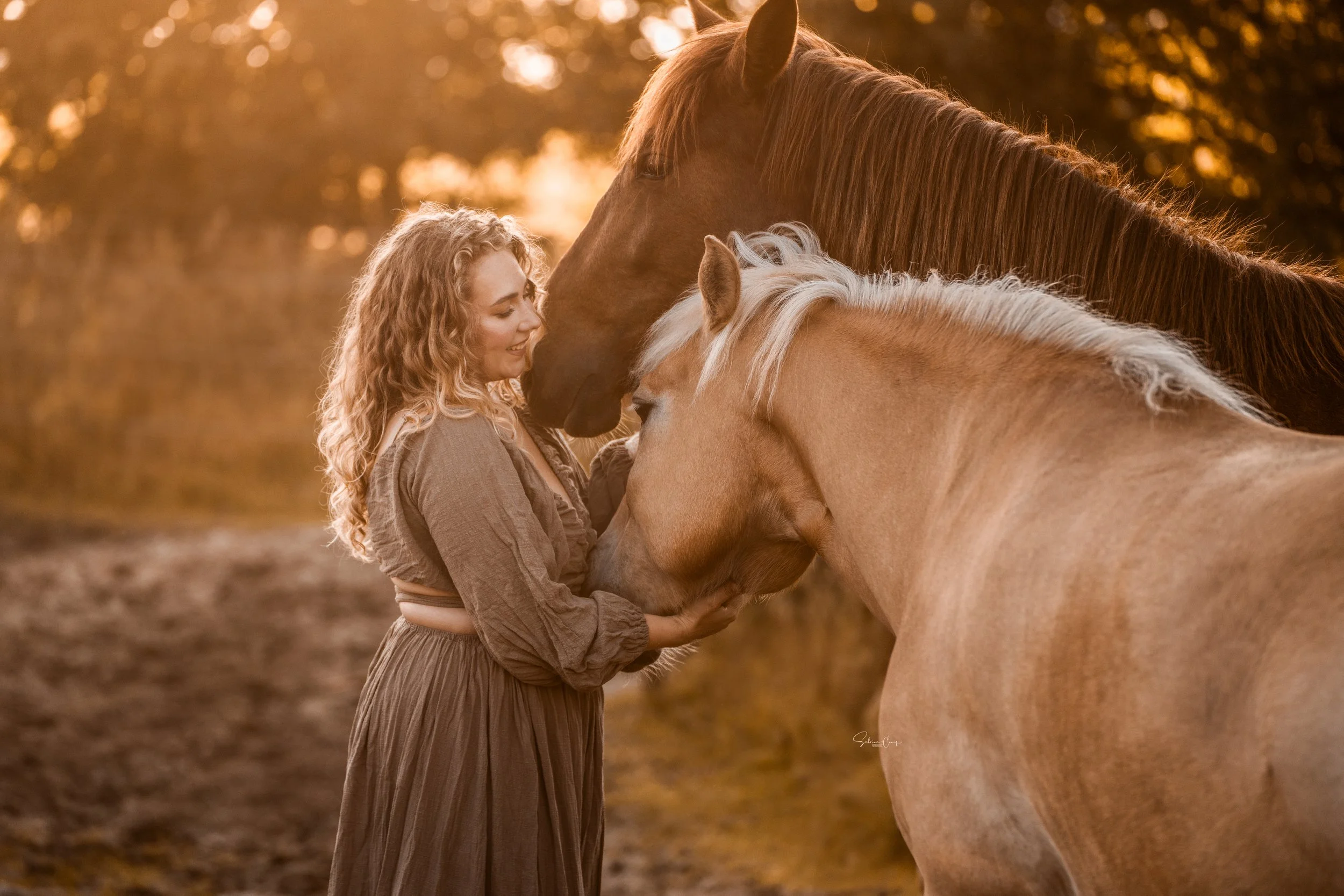 Eine Frau mit lockigem blondem Haar, die lächelt und zwei Pferde umarmt – ein braunes und ein hellbraunes – in einem Wald bei Sonnenuntergang.