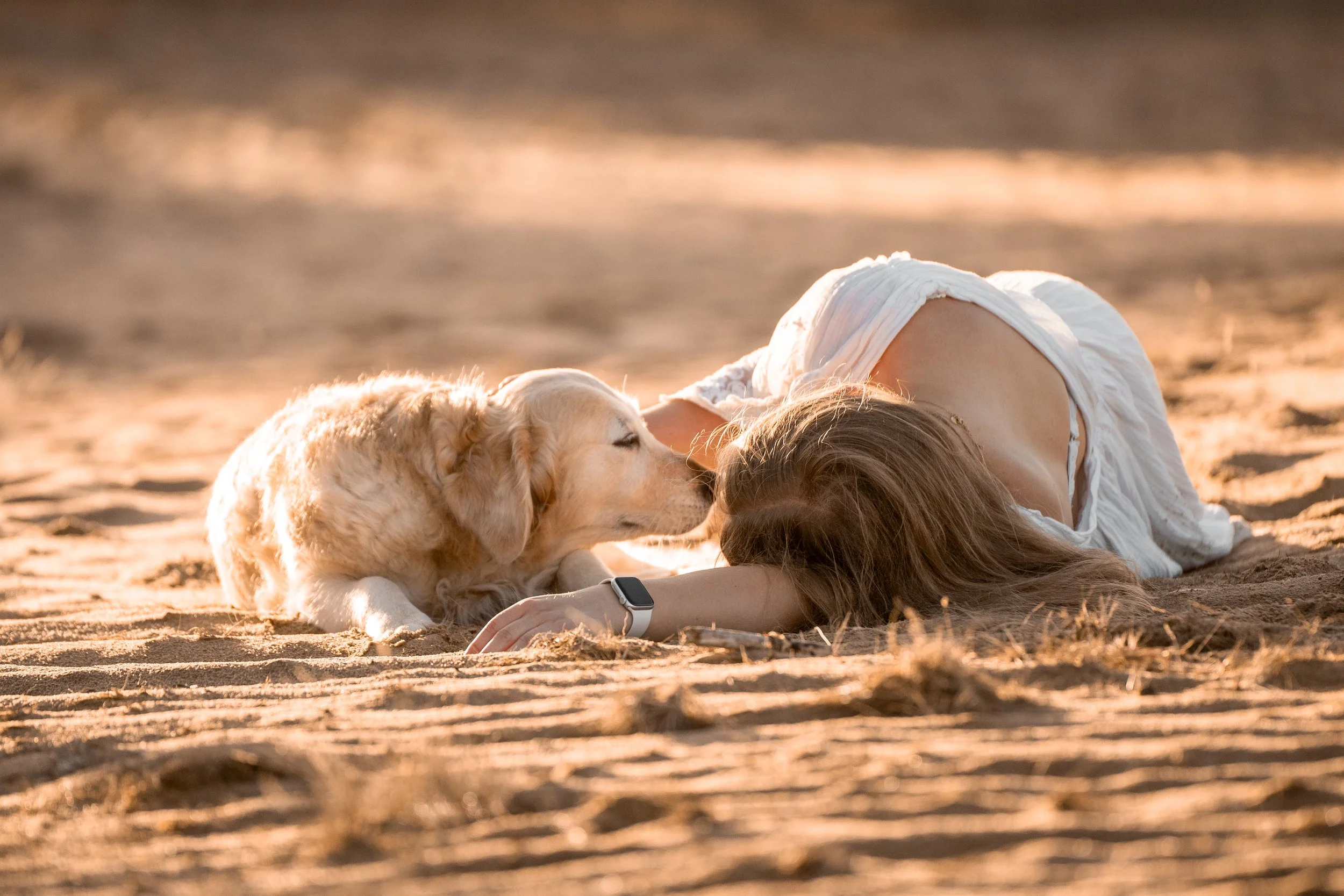 A woman lying on the sandy beach, resting her head on a dog, both soaking in the warm glow of sunset.