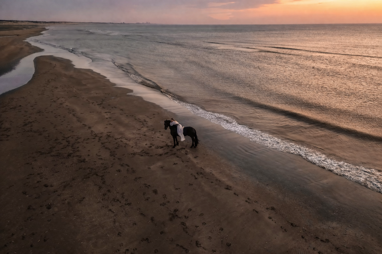 Ein Reiter auf einem schwarzen Pferd steht bei Sonnenuntergang an einem Strand, umgeben von Fußspuren im Sand und Wellen im Meer.