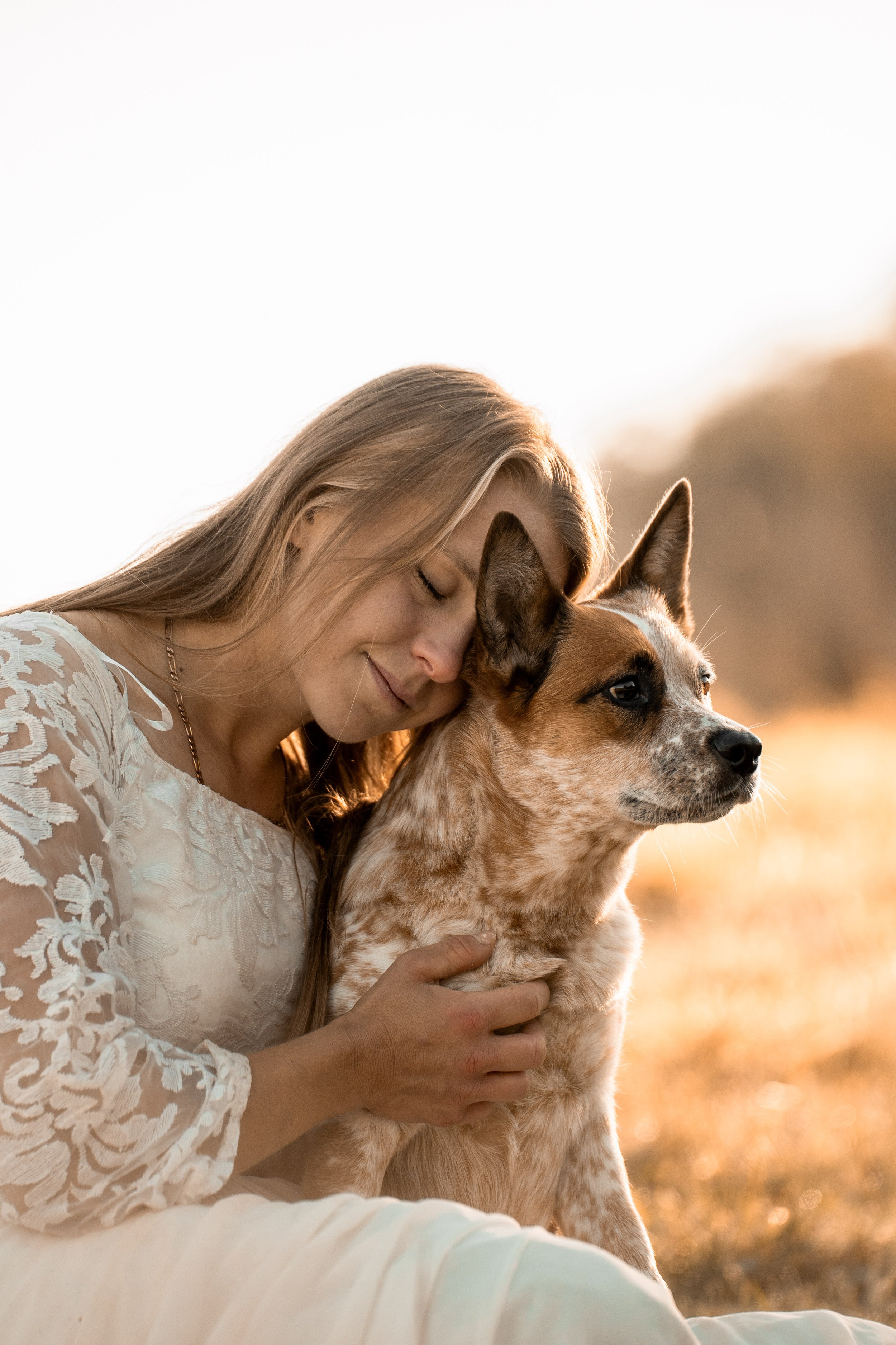 A woman with long blond hair and a white lace dress is hugging a brown and white dog outdoors during sunset.