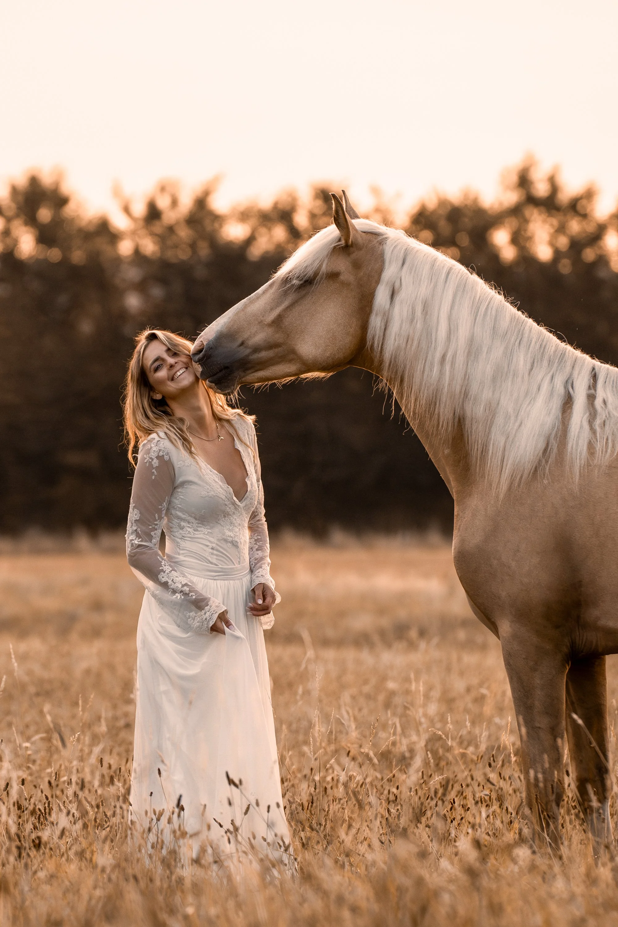 Eine Frau in einem weißen Kleid steht lächelnd in einem Feld mit hohem Gras und hat ihr Gesicht bei Sonnenuntergang dicht an die Nase eines großen, hellfarbigen Pferdes geschmiegt.