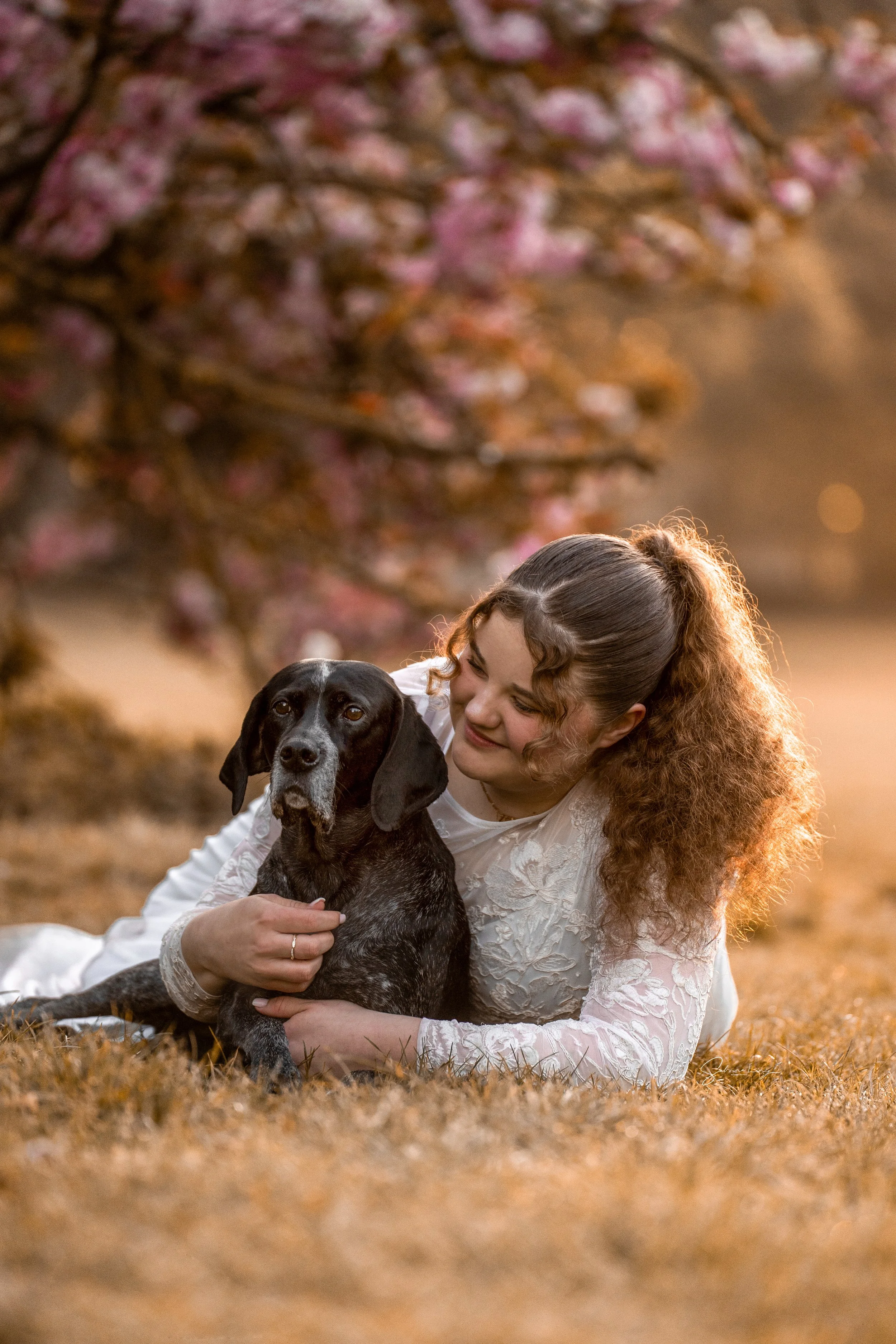 A woman with curly red hair lying on the grass, hugging a black and brown dog, with blurred pink blossoms on a tree in the background during golden hour.