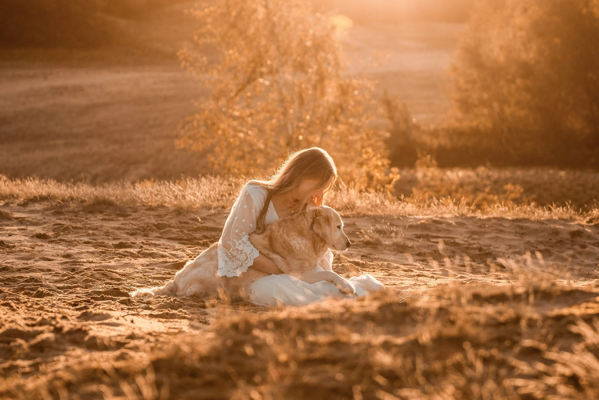 A young girl with long blonde hair sitting on the ground outdoors during sunset, hugging a light-colored dog, with sunlight creating a warm glow and a blurred background of trees.