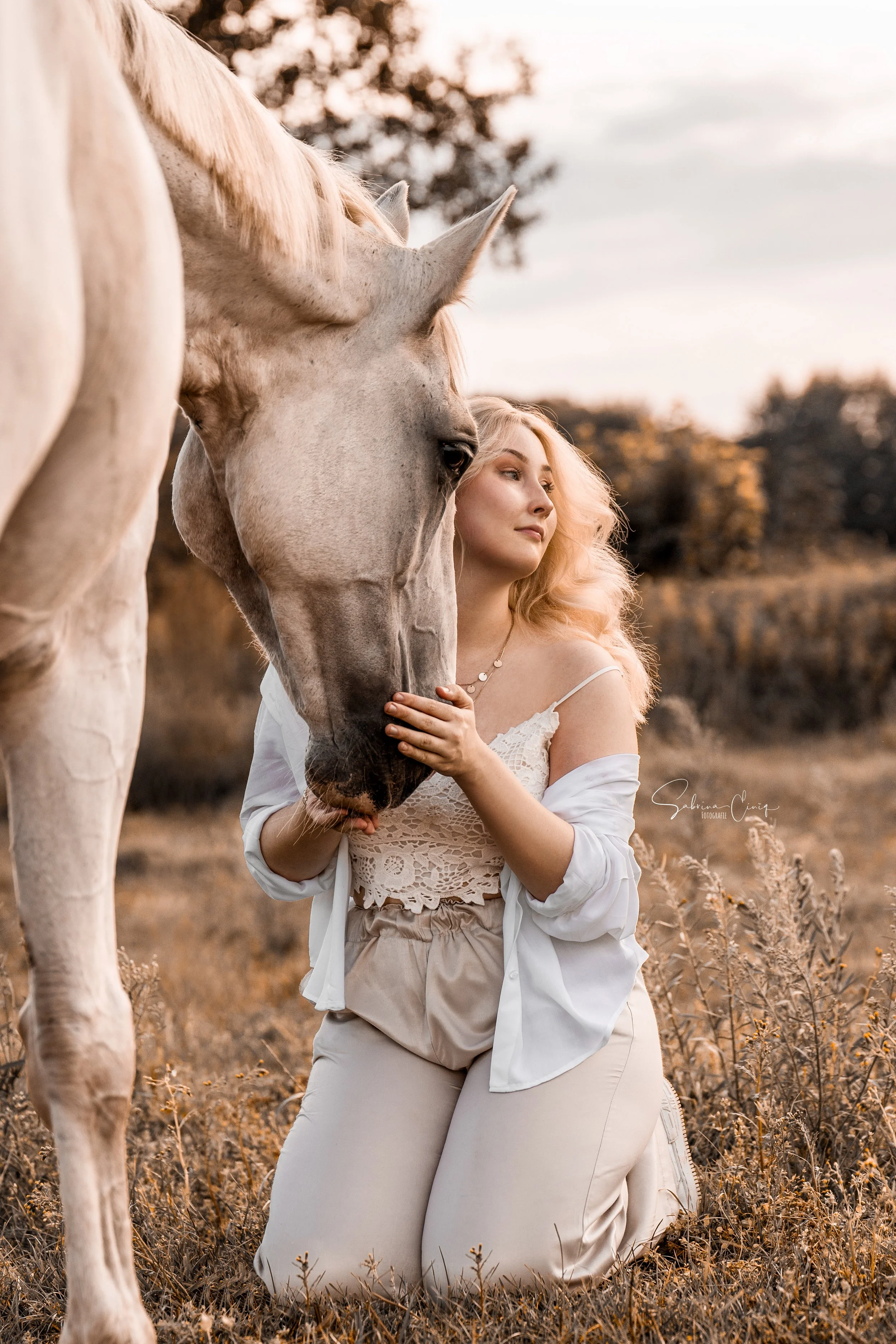 Eine Frau mit langen blonden Haaren, die eine weiße Bluse und eine beige Hose trägt, kniet auf trockenem Gras und hält das Gesicht eines großen weißen Pferdes in einem Feld bei Sonnenuntergang.