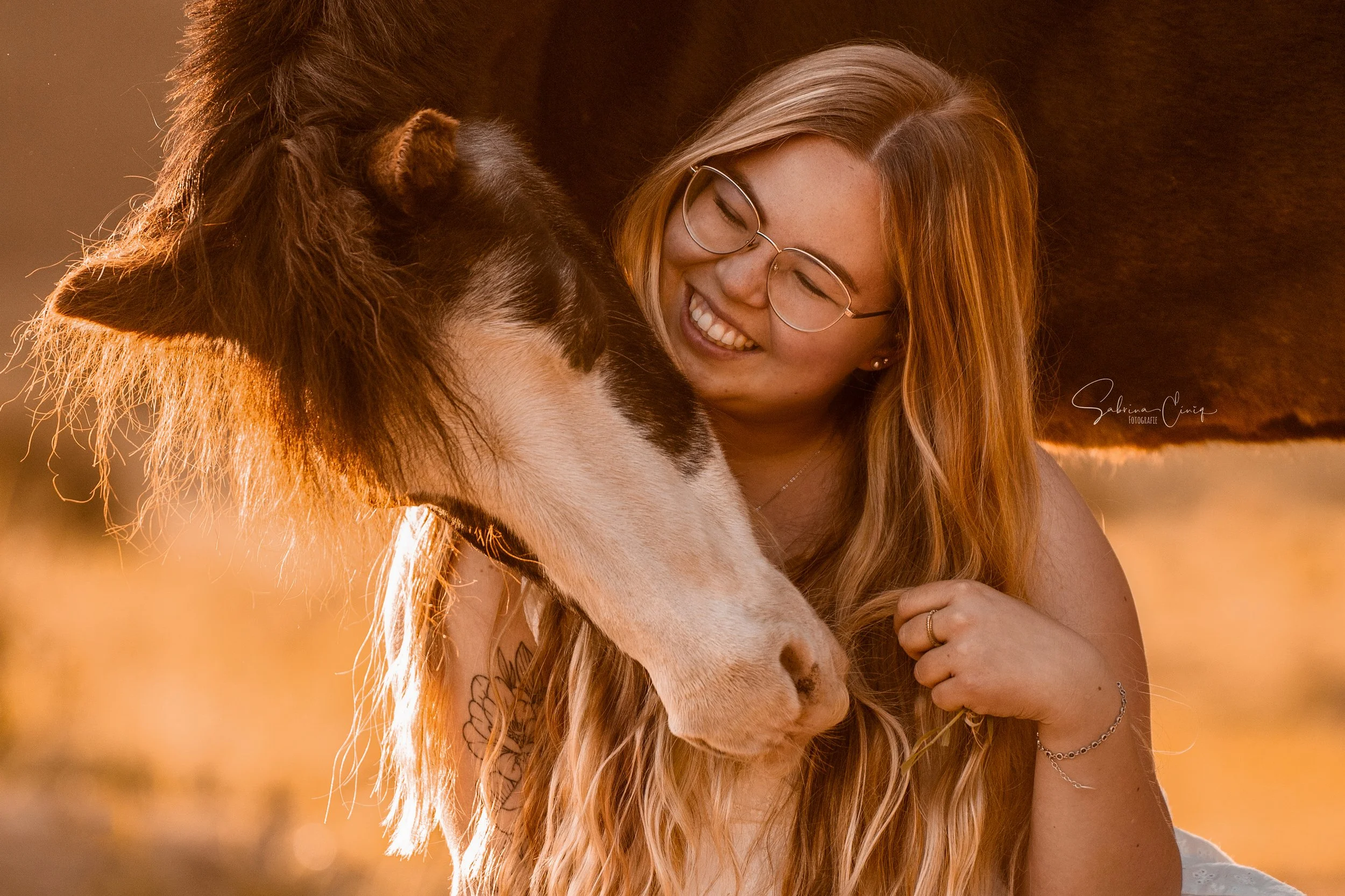 Eine Frau mit langen roten Haaren und Brille, die lächelt, während sie bei Sonnenuntergang ein Pferd auf einer Wiese umarmt.