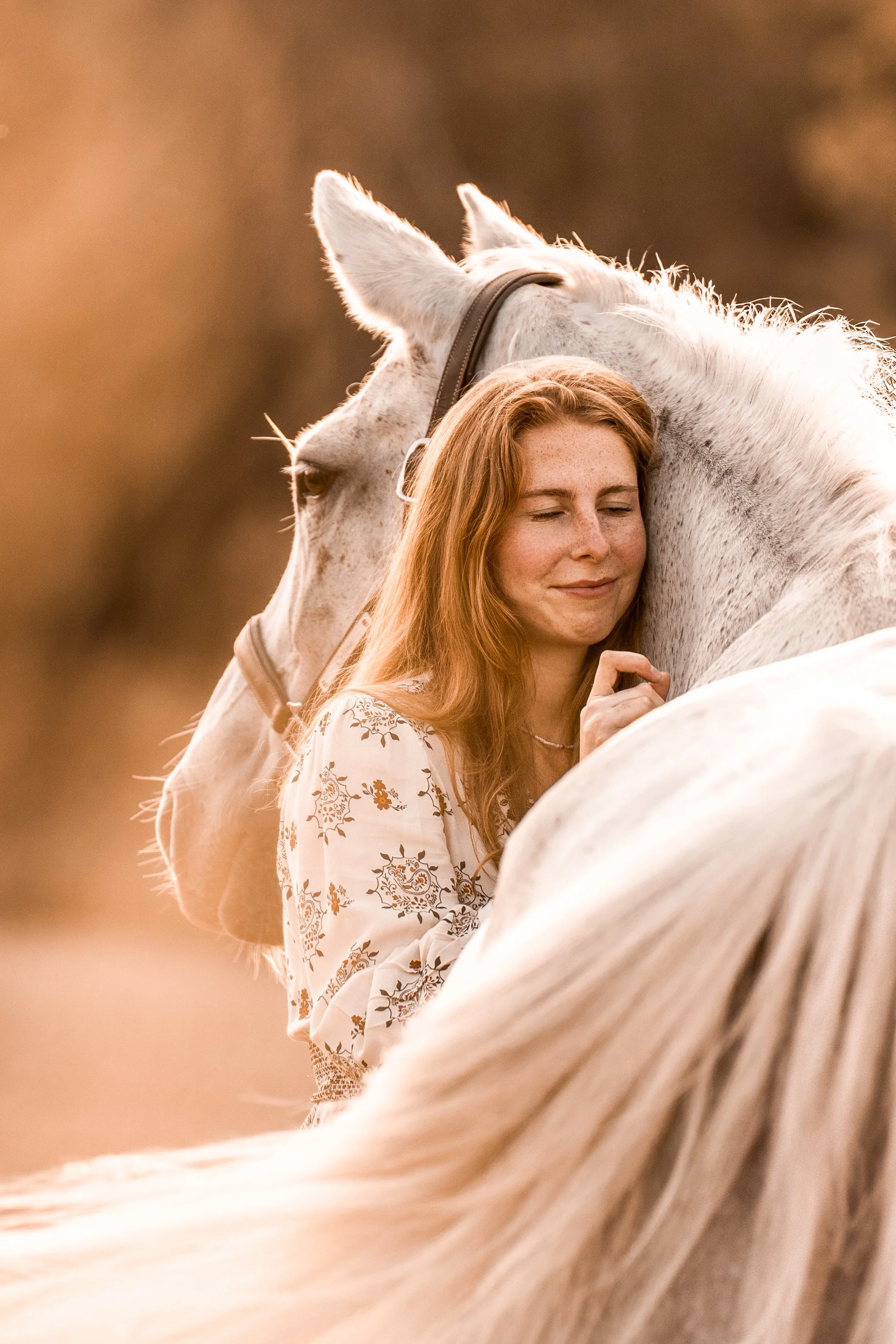 Eine Frau mit roten Haaren und Sommersprossen umarmt ein weißes Pferd; beide haben die Augen geschlossen, während die Sonne untergeht oder die „goldene Stunde“ anbricht.