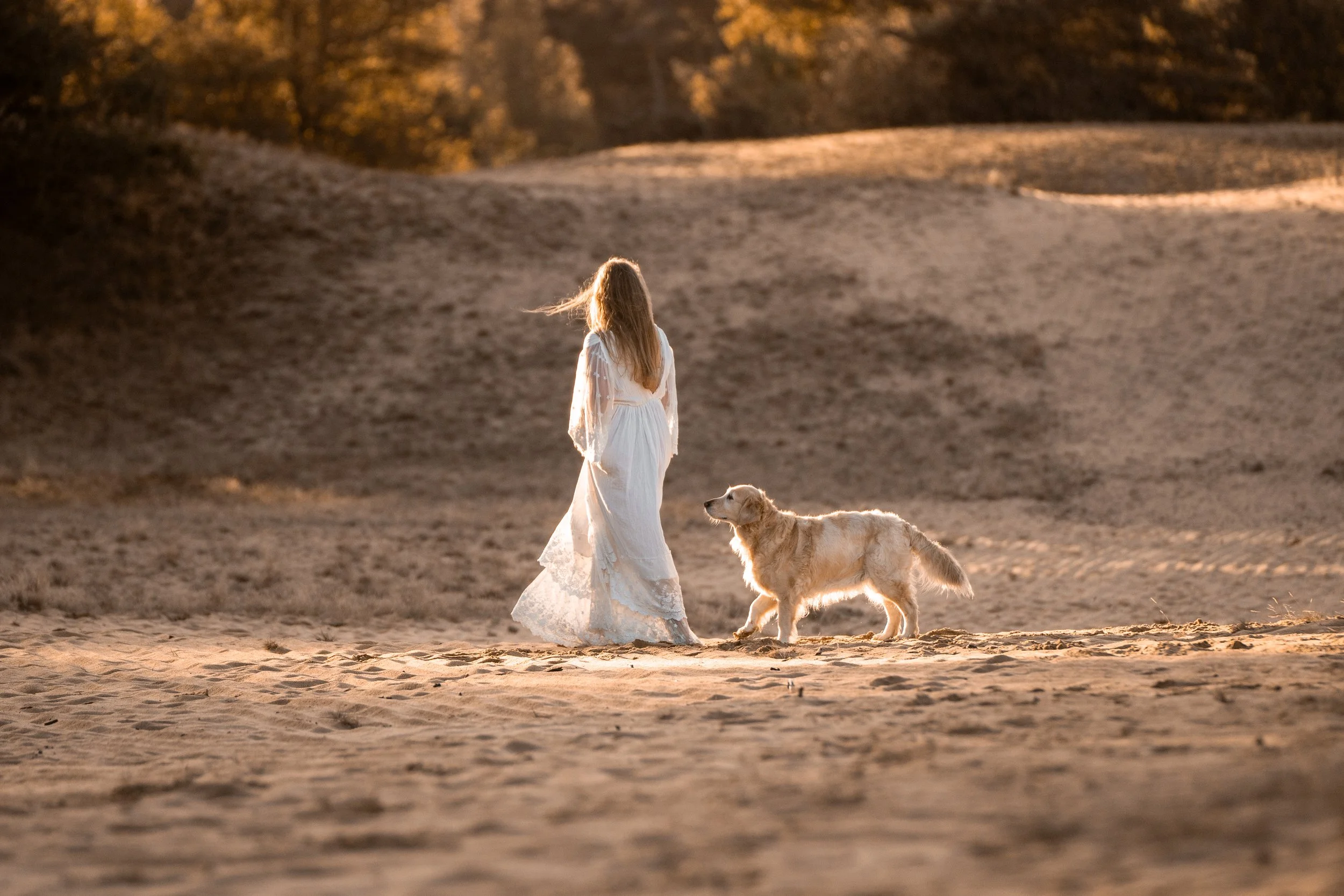 A woman in a white dress walking with a dog on a sandy landscape during sunset or sunrise with warm lighting and trees in the background.