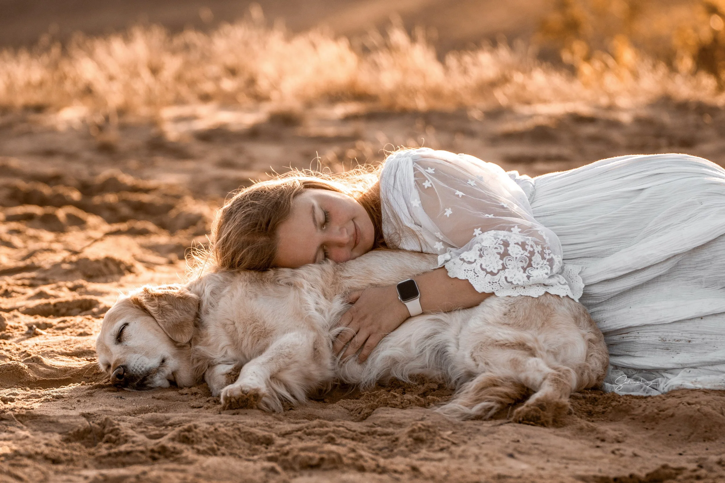 A woman with red hair lying on the ground in a white dress, hugging a golden retriever puppy, both sleeping peacefully on a sandy area with warm sunset lighting.