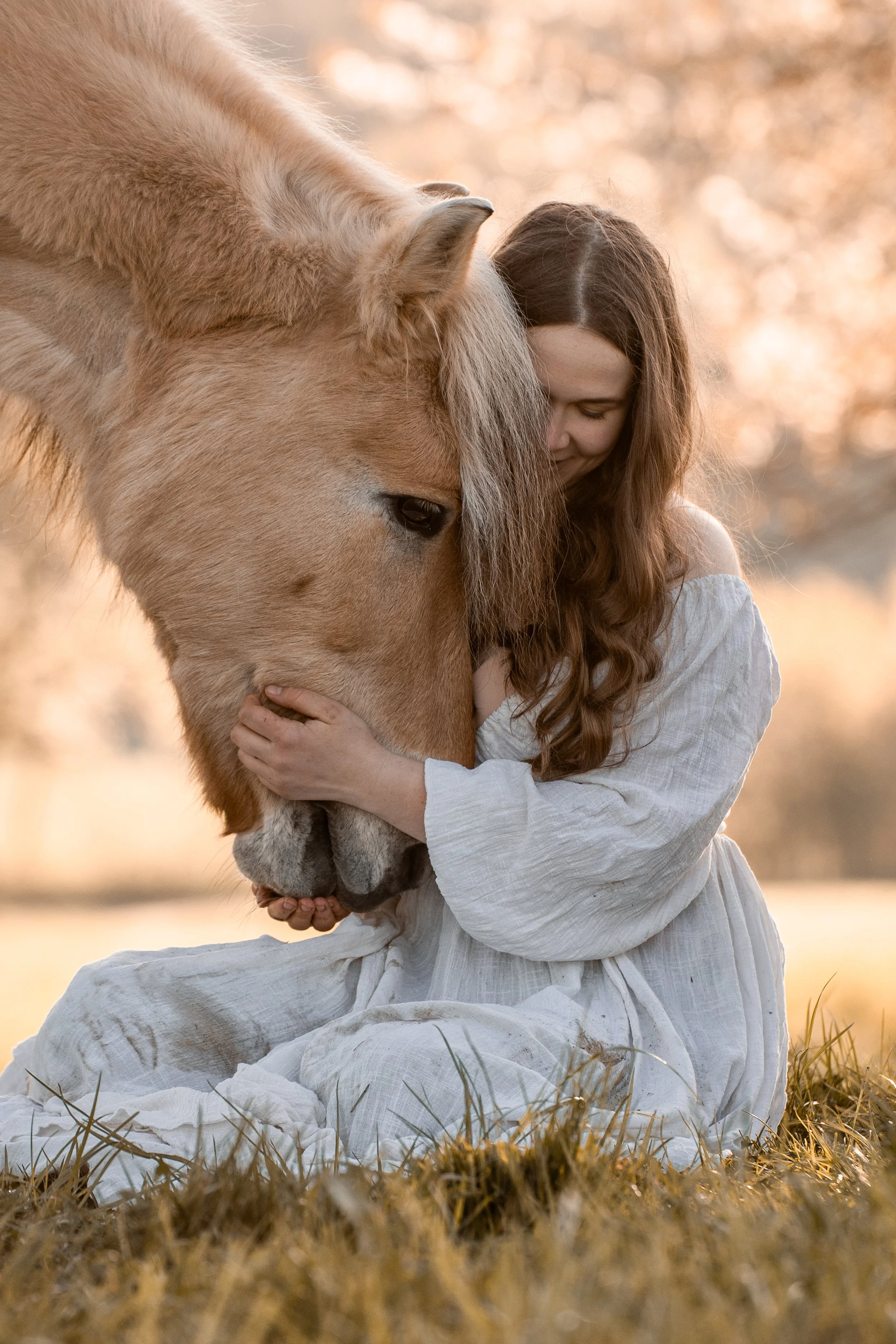Eine junge Frau mit langen braunen Haaren, die ein weißes Kleid trägt, sitzt im Gras und umarmt bei Sonnenuntergang ein helles Pferd.