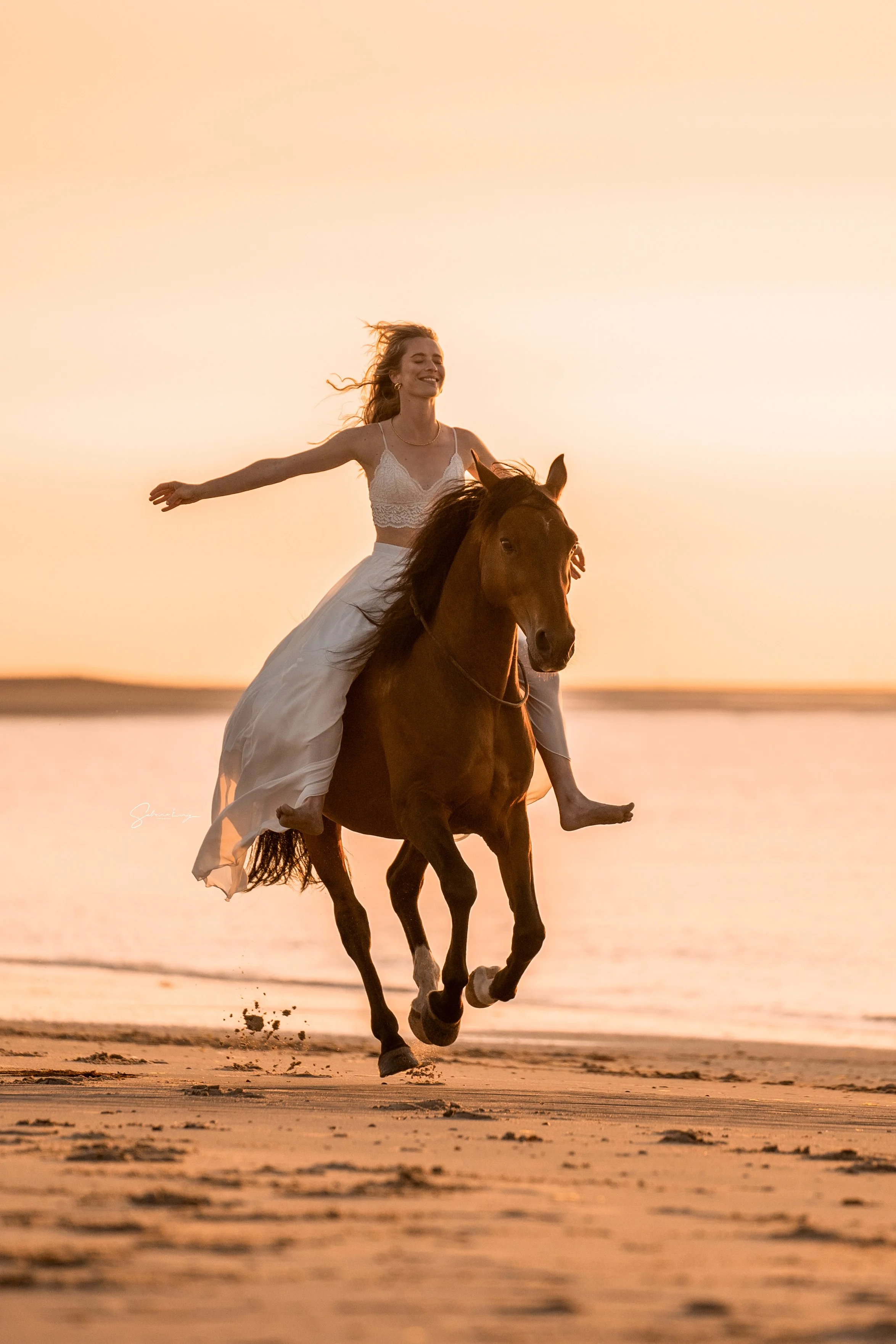 Eine Frau in einem weißen Kleid reitet bei Sonnenuntergang auf einem galoppierenden braunen Pferd am Strand entlang.
