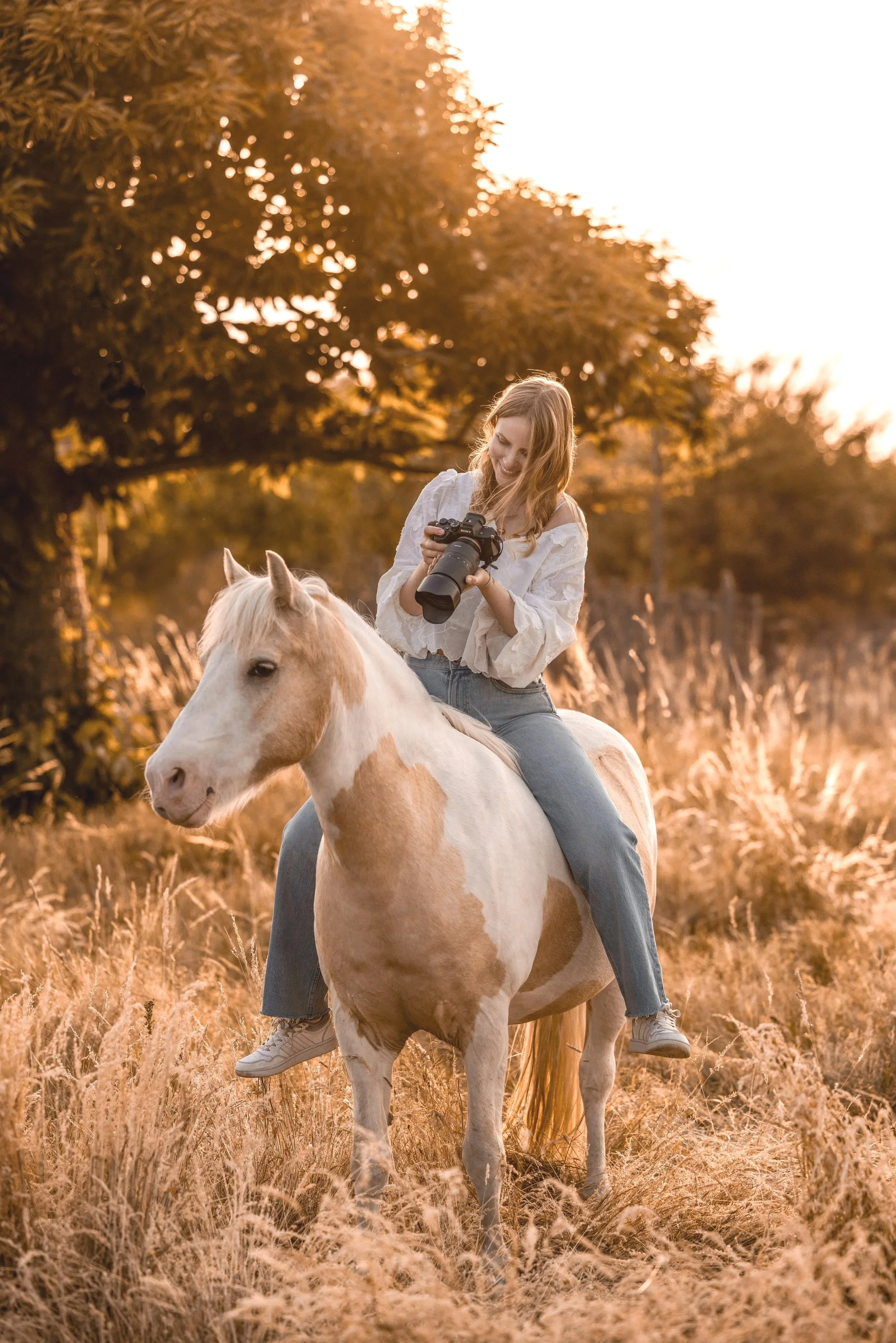 Eine junge Frau reitet bei Sonnenuntergang auf einem weißen Pferd durch eine Wiese, umgeben von Bäumen mit goldenem Laub.