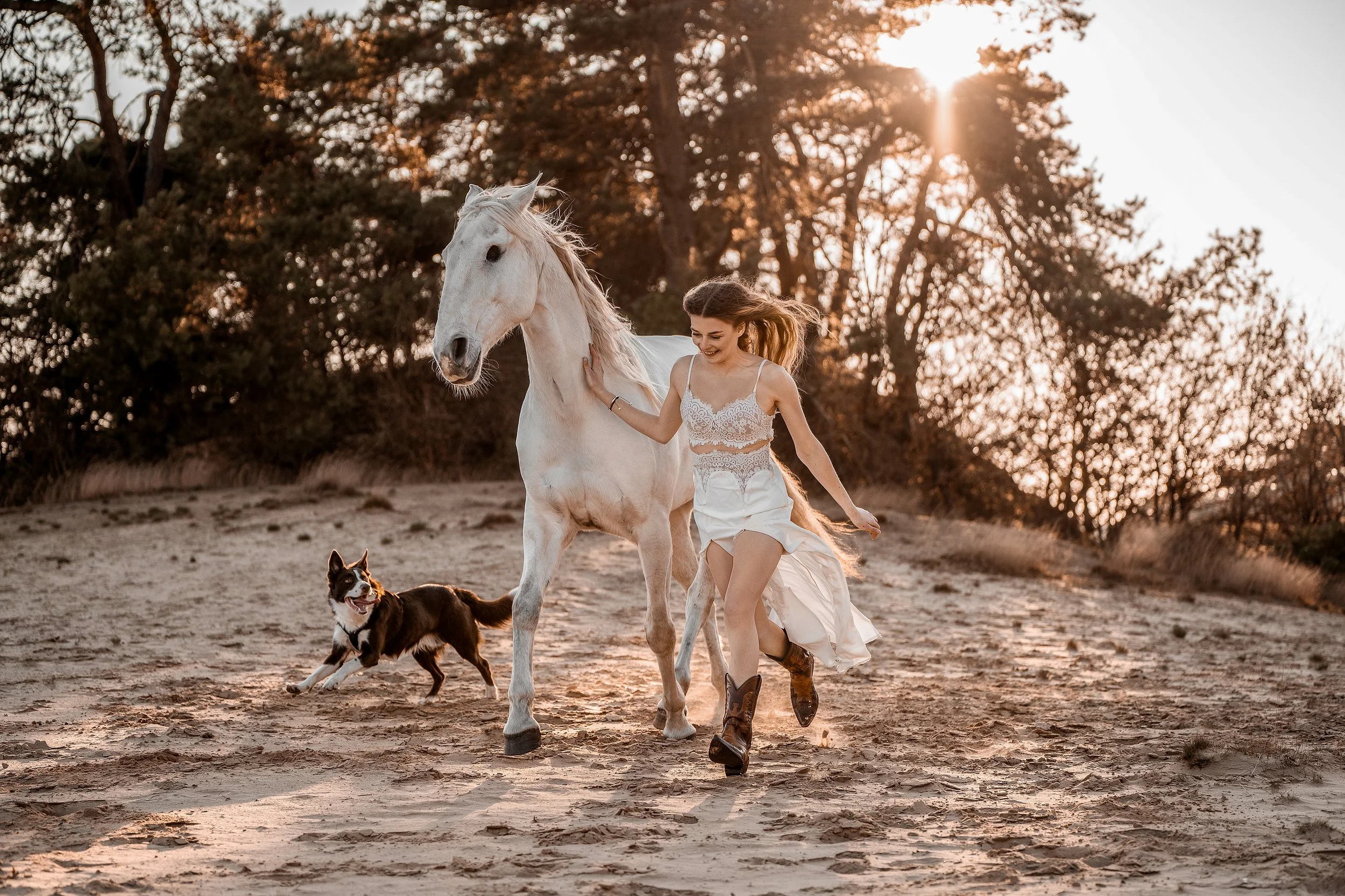 Eine junge Frau in einem weißen Kleid, die bei Sonnenuntergang mit einem weißen Pferd und zwei Hunden auf einem Feldweg spazieren geht.