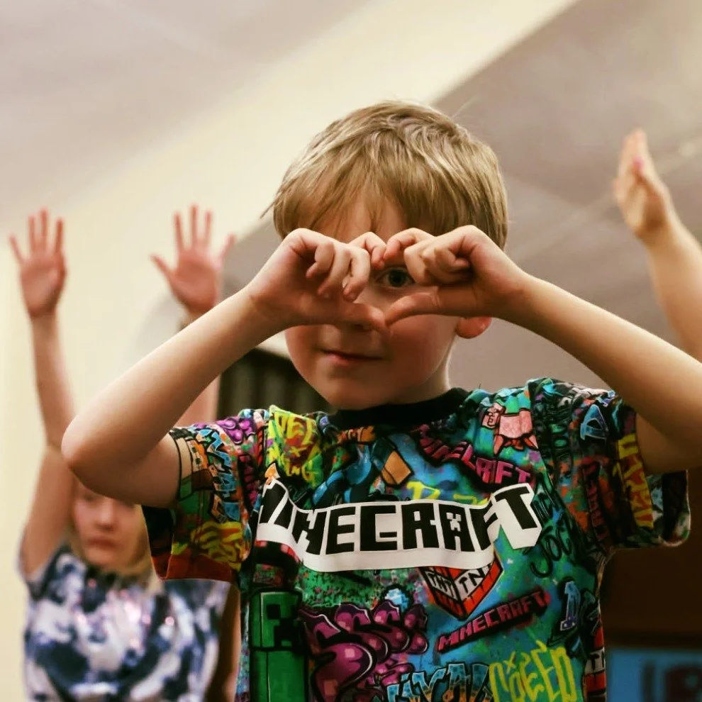 A young boy with light brown hair making a heart shape with his hands over his face, wearing a colorful Minecraft-themed shirt. Two other children are seen in the background with their hands raised.