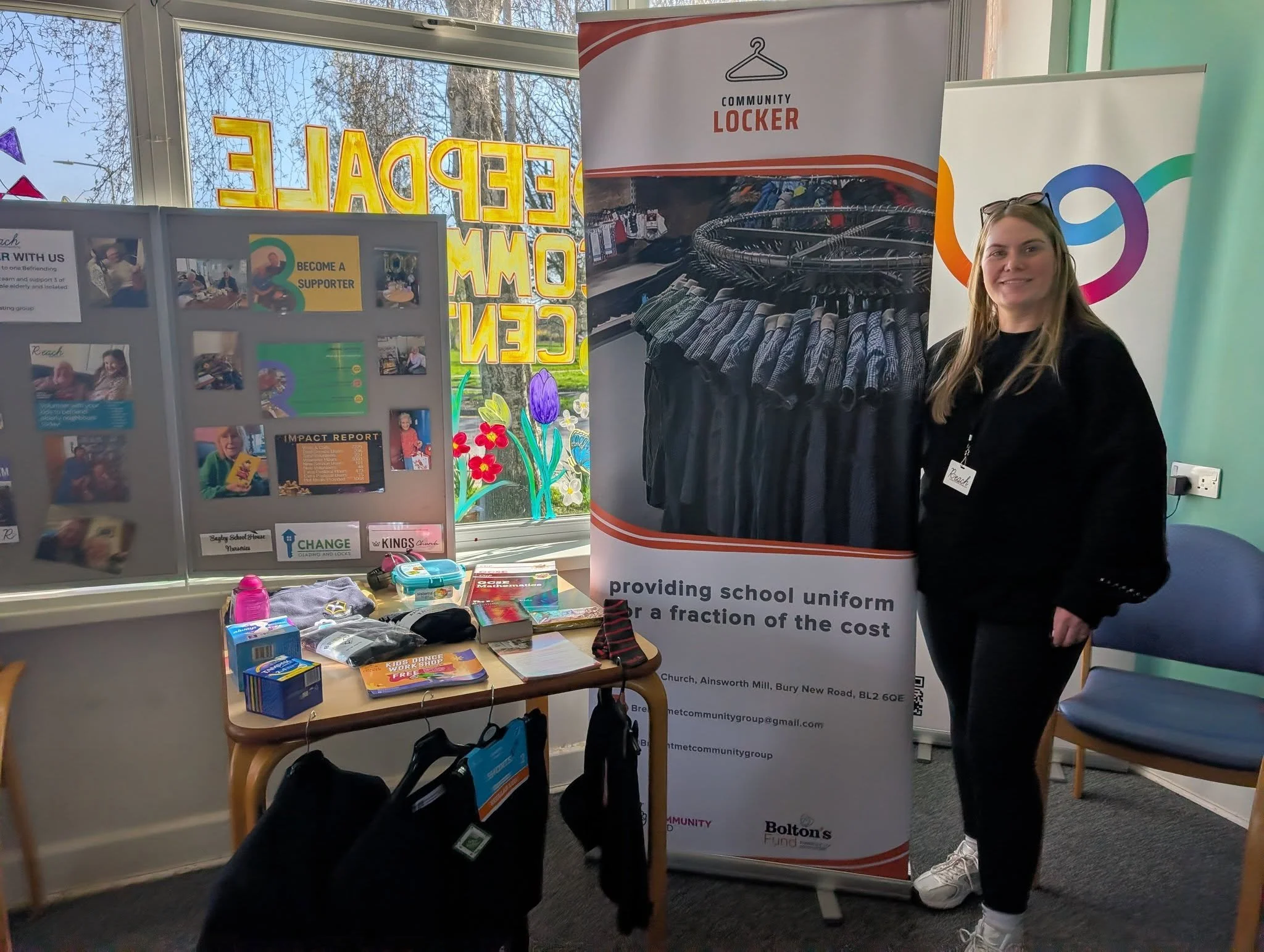 A young woman standing next to a promotional banner for providing school uniforms at a community event. The banner shows a photo of black school uniforms on hangers. There is a table with various items, including books, water bottles, and bags, and a display board behind with photos and messages about supporting children. The window behind has colorful decorations and the word 'PEACE' spelled out in large, yellow letters.