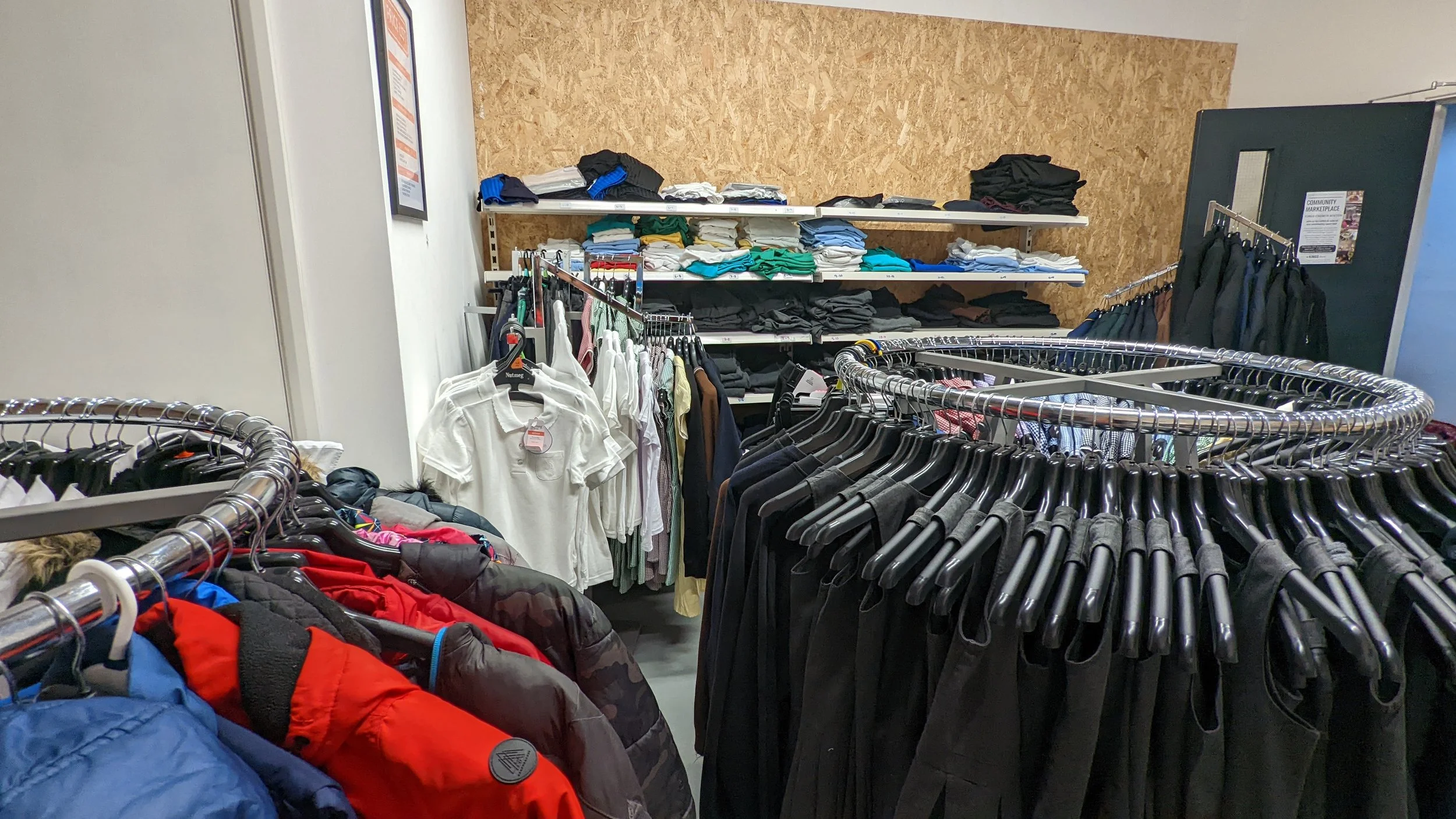 Clothing racks with jackets and shirts in a thrift store, shelves with folded jeans and t-shirts in the background.