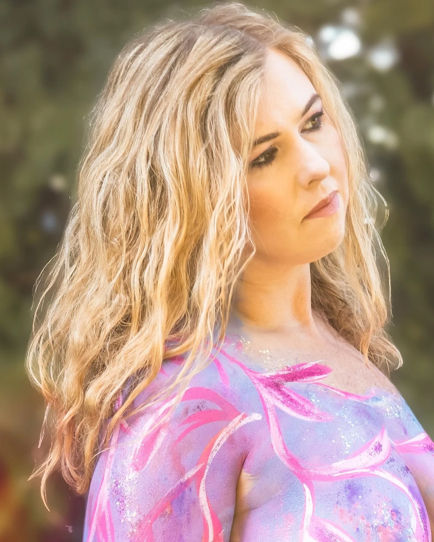 A woman with wavy blonde hair in a colorful, painted or body-painted outfit outdoors, looking to the side.