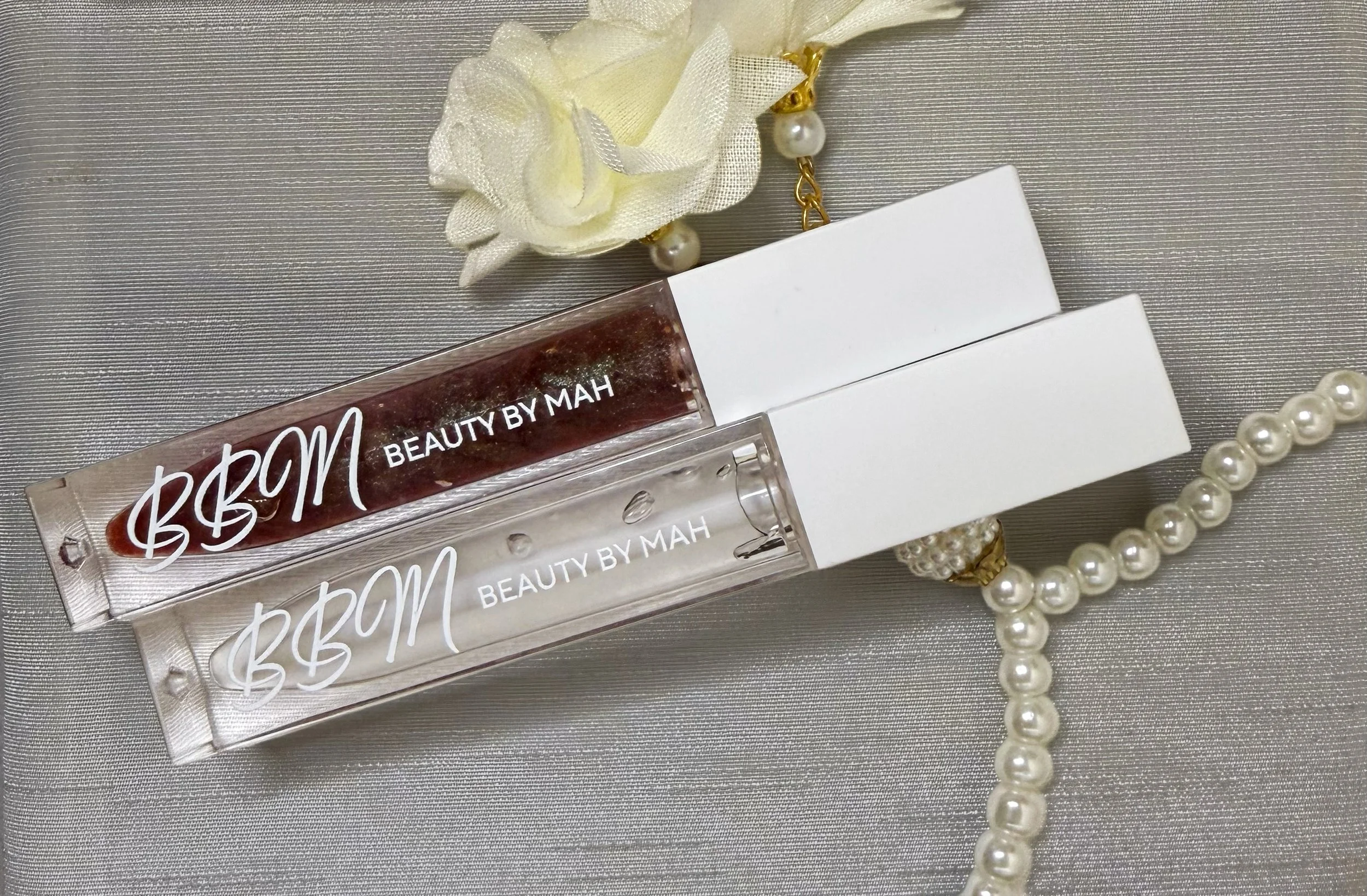 Two lip gloss tubes labeled 'Beauty by Mah', one with dark red gloss and the other with clear gloss, placed on a satin fabric surface. Accompanying accessories include a string of pearl beads and a fabric flower with pearl earrings.