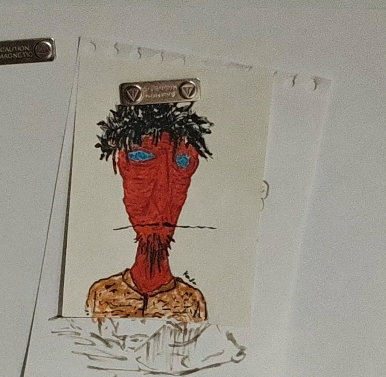 A hand-drawn portrait of a person with red skin, black spiky hair, blue eyes, and a beard, wearing a beige shirt, taped to a white wall.