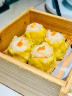 A wooden steamer basket containing four pieces of shrimp dumplings with yellow wrappers and white topping, garnished with small orange bits.