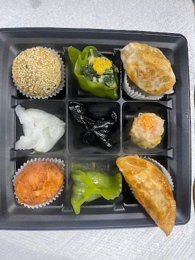 A black tray with nine compartments containing assorted dim sum, including sesame-covered bun, green pepper with yellow filling, fried dumpling, white dumpling, black sesame ball, shrimp dumpling, orange crab puff, green chili, and fried vegetable ro