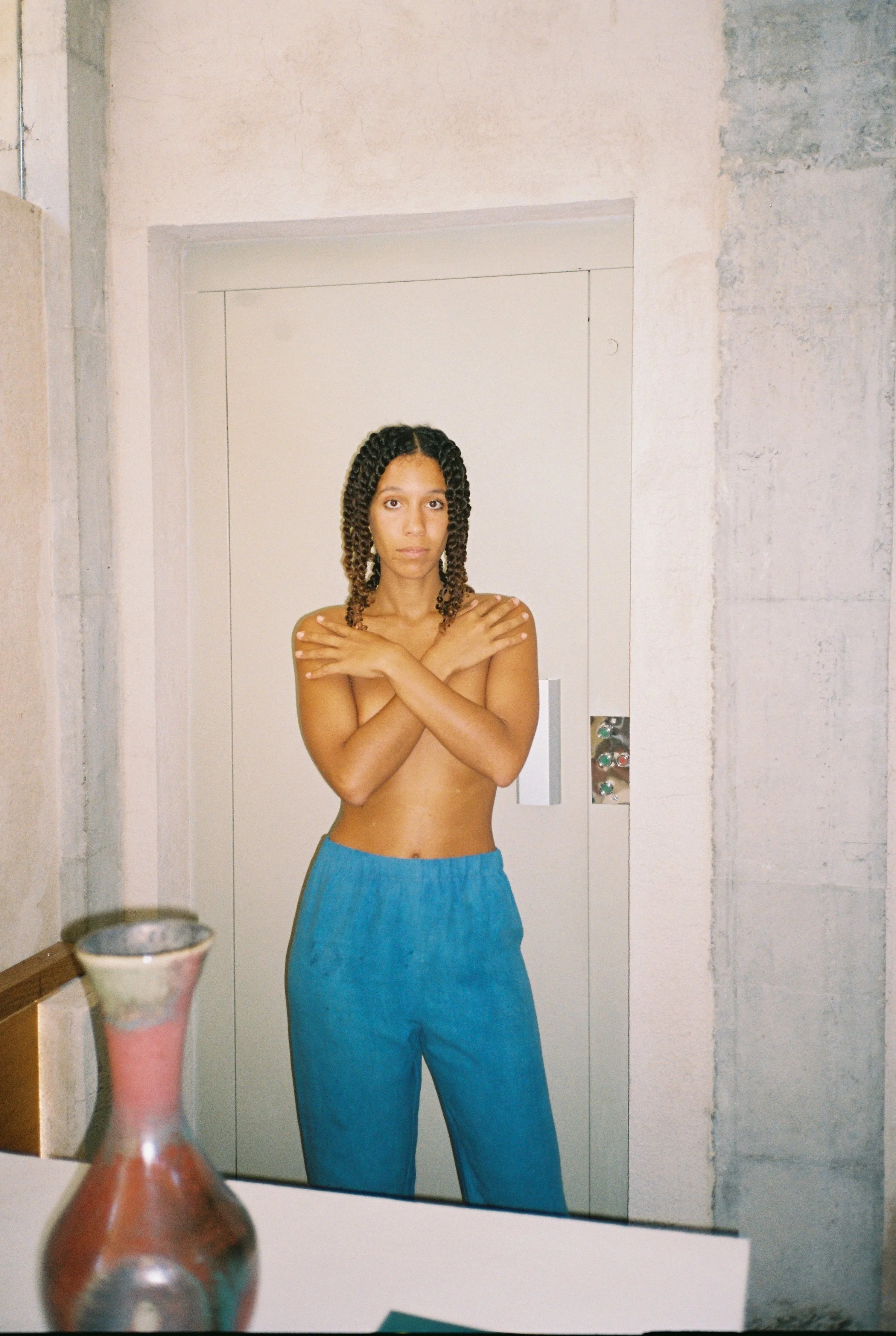A woman with curly hair stands topless in front of a white door, wearing blue pants and crossing her arms over her shoulders. There is a vase on the table in the foreground.