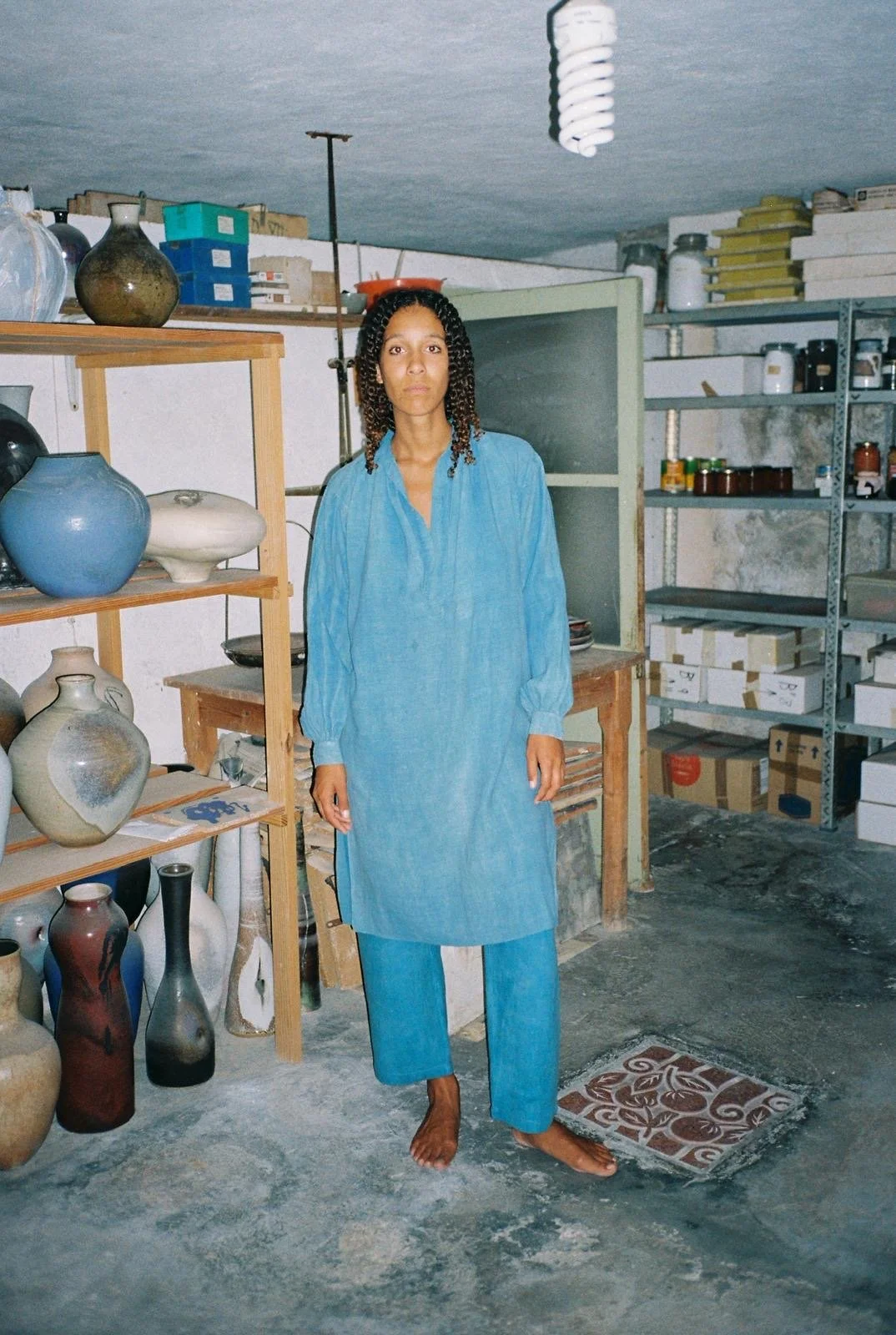A woman in a blue dress and matching pants standing barefoot in a ceramics studio surrounded by shelves of pottery and storage boxes.