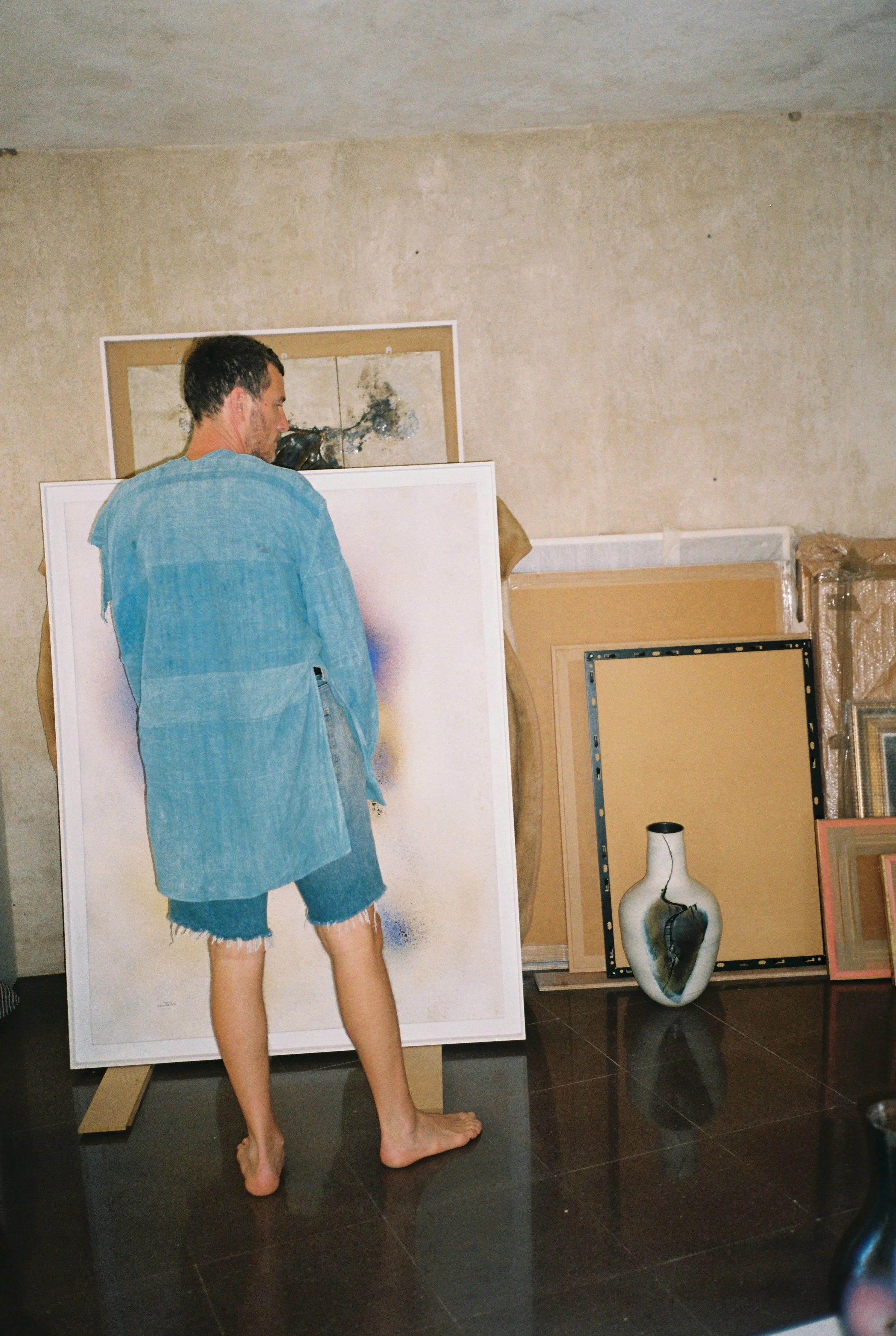 An artist stands barefoot in a studio filled with paintings and artwork, observing a large abstract painting.