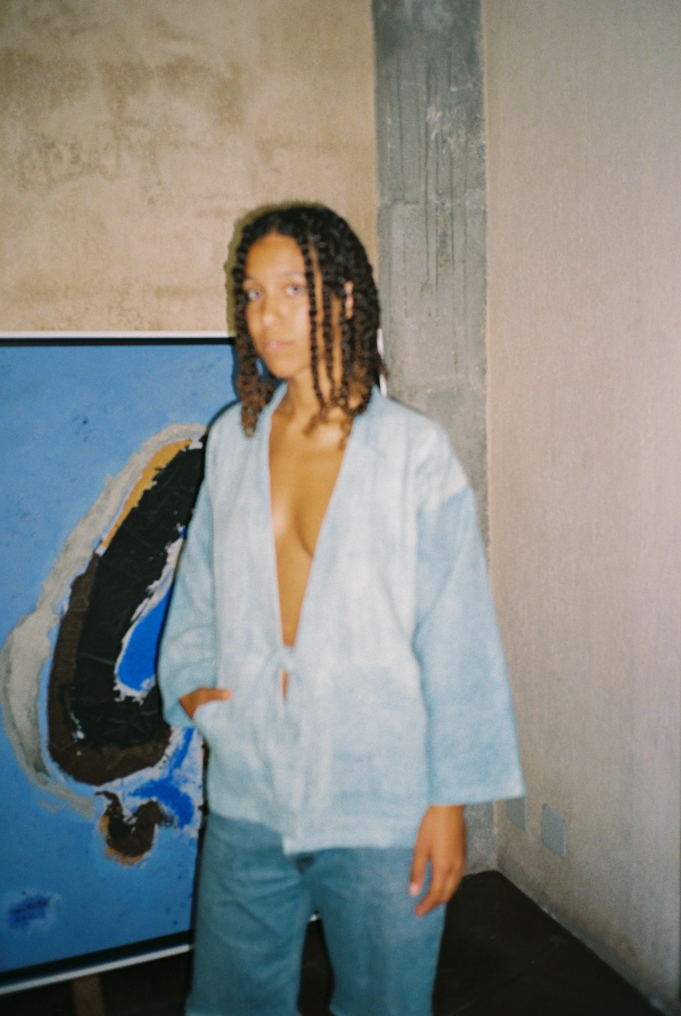 A woman with braided hair wearing a light denim blazer and matching jeans, standing in front of a colorful abstract painting and a textured wall.