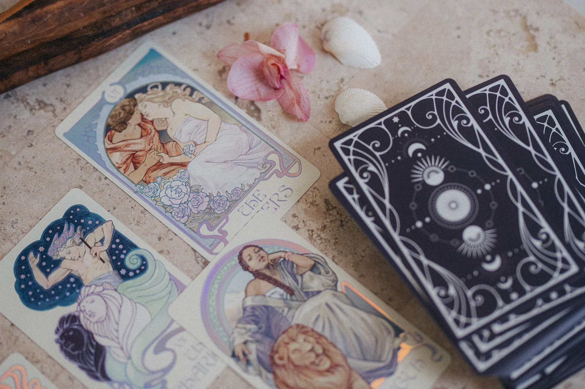 A tarot card reading setup with multiple tarot cards laid out on a table, including a card with an illustration of two women, and a deck of black-backed tarot cards nearby. There are also pink and white seashells and pink flower petals around the car