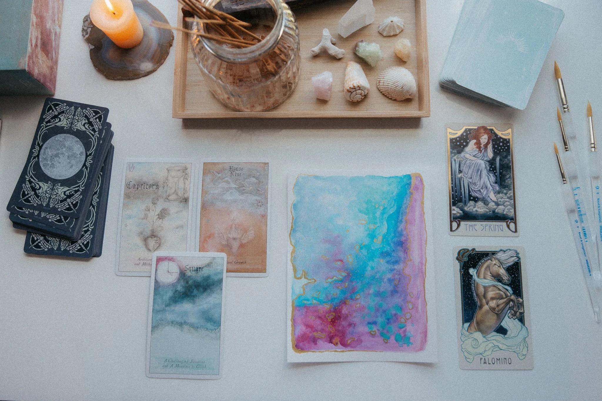 A tabletop setup for tarot or oracle card reading, featuring a deck of cards with intricate back designs, a tarot card illustrating a woman and a horse labeled 'Palomino,' several small cards with watercolor artwork and text, a colorful picturesque map, a jar with incense sticks, a stack of light blue cards, and a lit candle on a wooden slab.