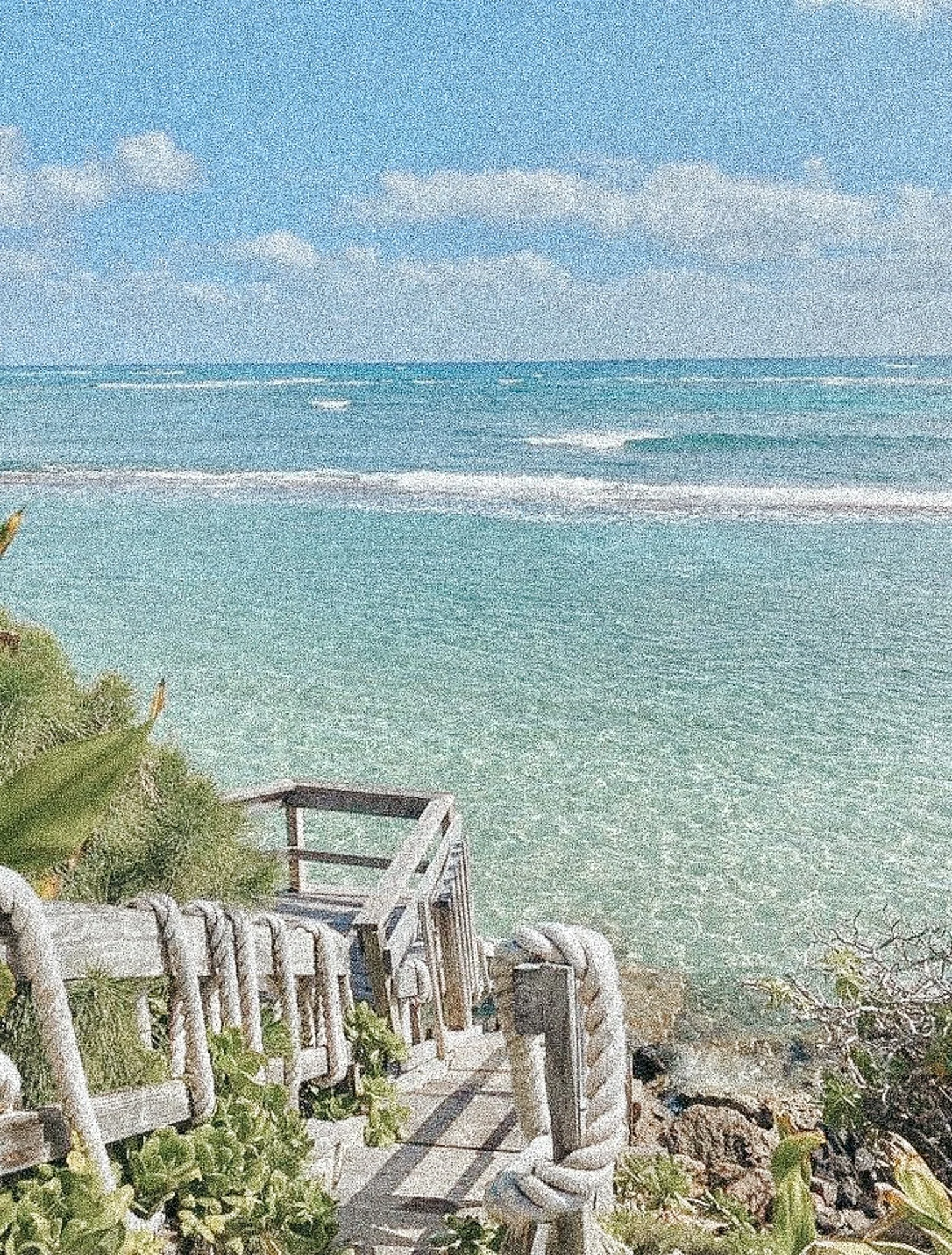 Wooden staircase leading down to a sandy beach with greenery on the sides, overlooking clear ocean water under a partly cloudy sky.
