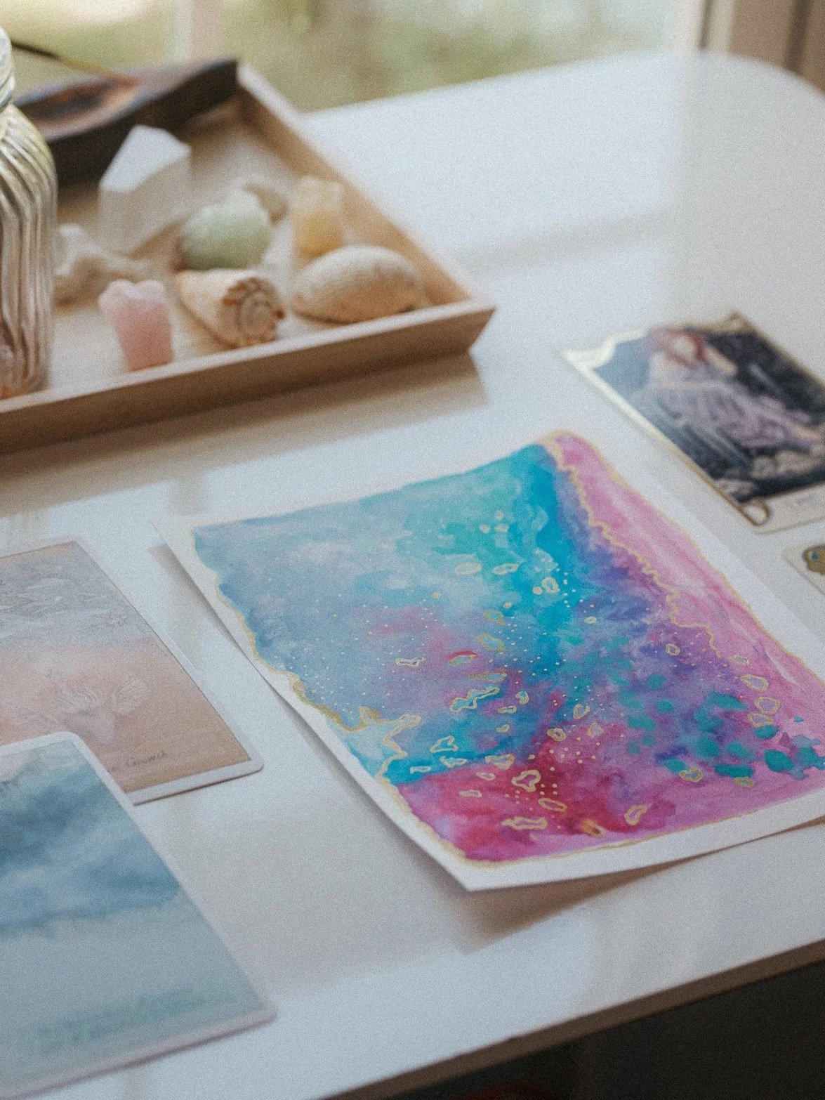 A table with watercolor art prints, a colorful abstract painting, and a tray of sea shells and small decorative objects.