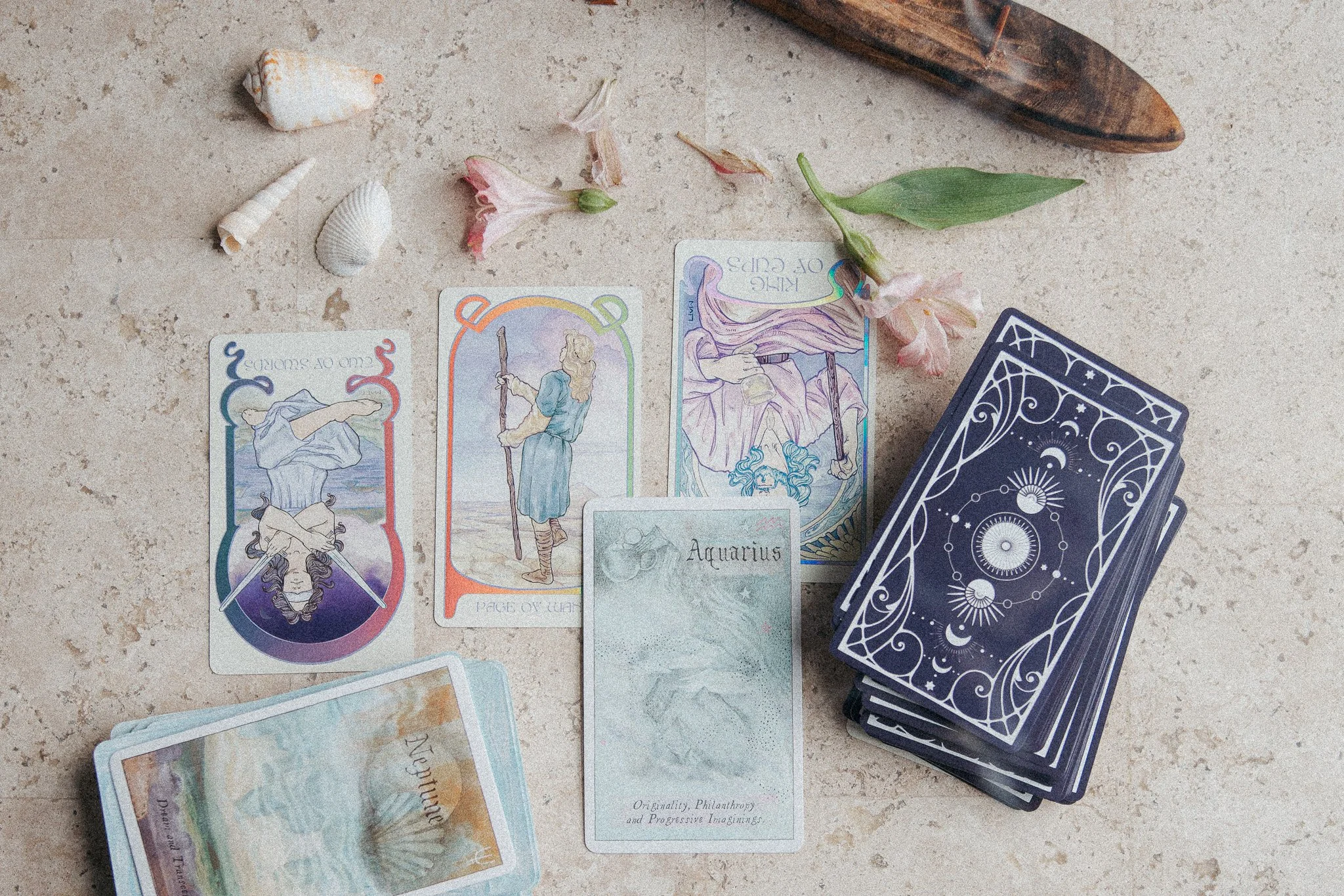 Tarot cards spread out on a surface with shells, flowers, a wooden object, and a deck of back-facing cards nearby.