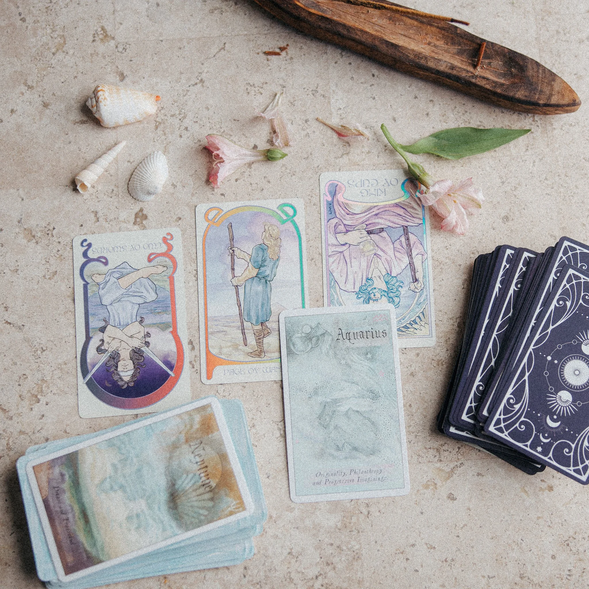 A spread of tarot cards and tarot card decks on a beige surface, with seashells, a flower, a green leaf, a wooden object, and some colorful illustrated documents.