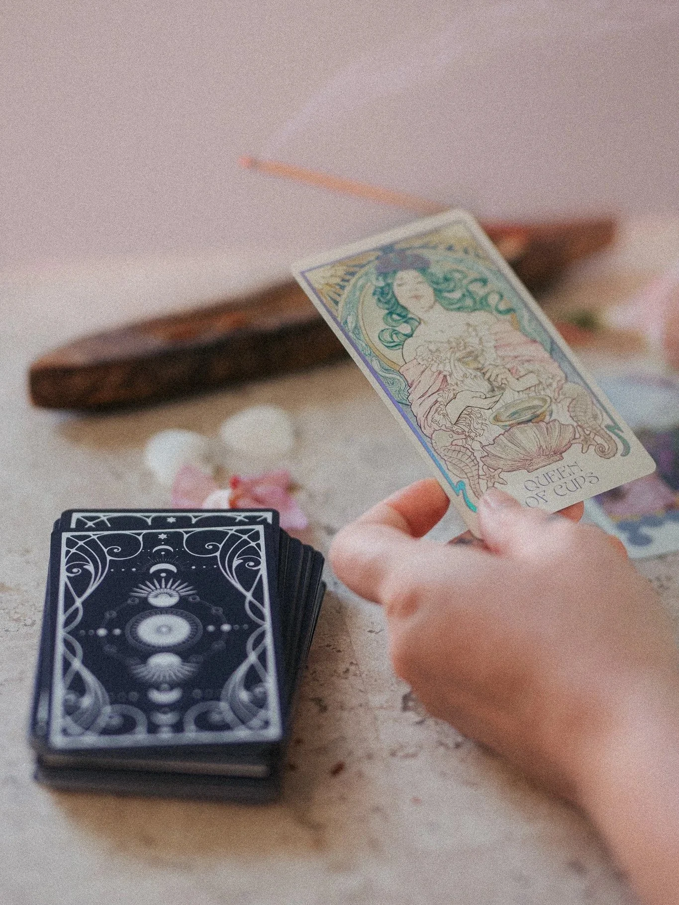 A tarot card reading scene with a hand holding a card titled "Queen of Cups," tarot cards with a black and white design on the table, and a deck of tarot cards face down nearby.