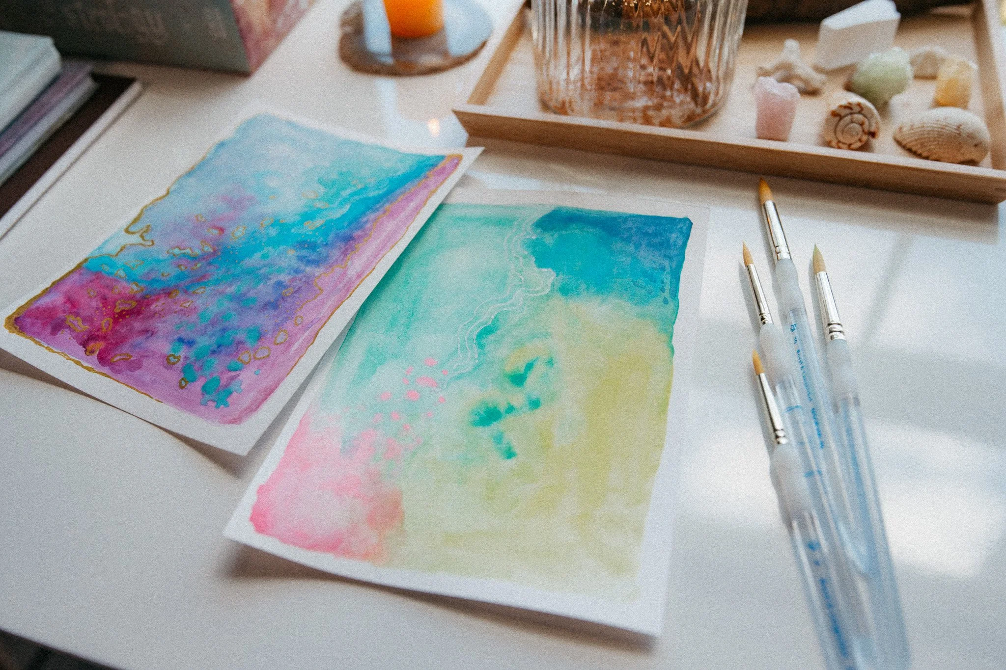 Two watercolor paintings with abstract designs in pink, blue, yellow, and green, placed on a white surface surrounded by art supplies including three paintbrushes, a glass jar filled with water, a wooden tray with seashells and small stones, and a stack of notebooks.