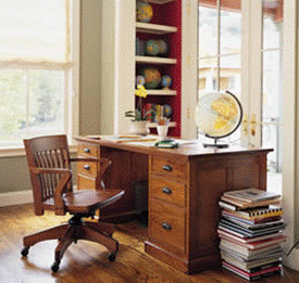 A wooden desk and chair in a home office with windows, a globe, and a shelf with books and decorative items.