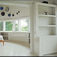 Living room with large windows, built-in white shelves, and a white wall. There are decorative plates on the wall near the ceiling and some greenery outside the window.