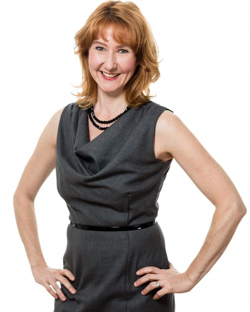 A woman with red hair, wearing a sleeveless gray dress and black necklace, posing with hands on hips and smiling.
