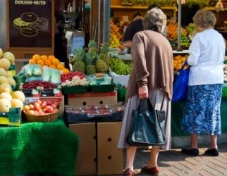 Two elderly women shopping at an outdoor fruit and vegetable stand with various fresh produce, including oranges, apples, and pineapples.