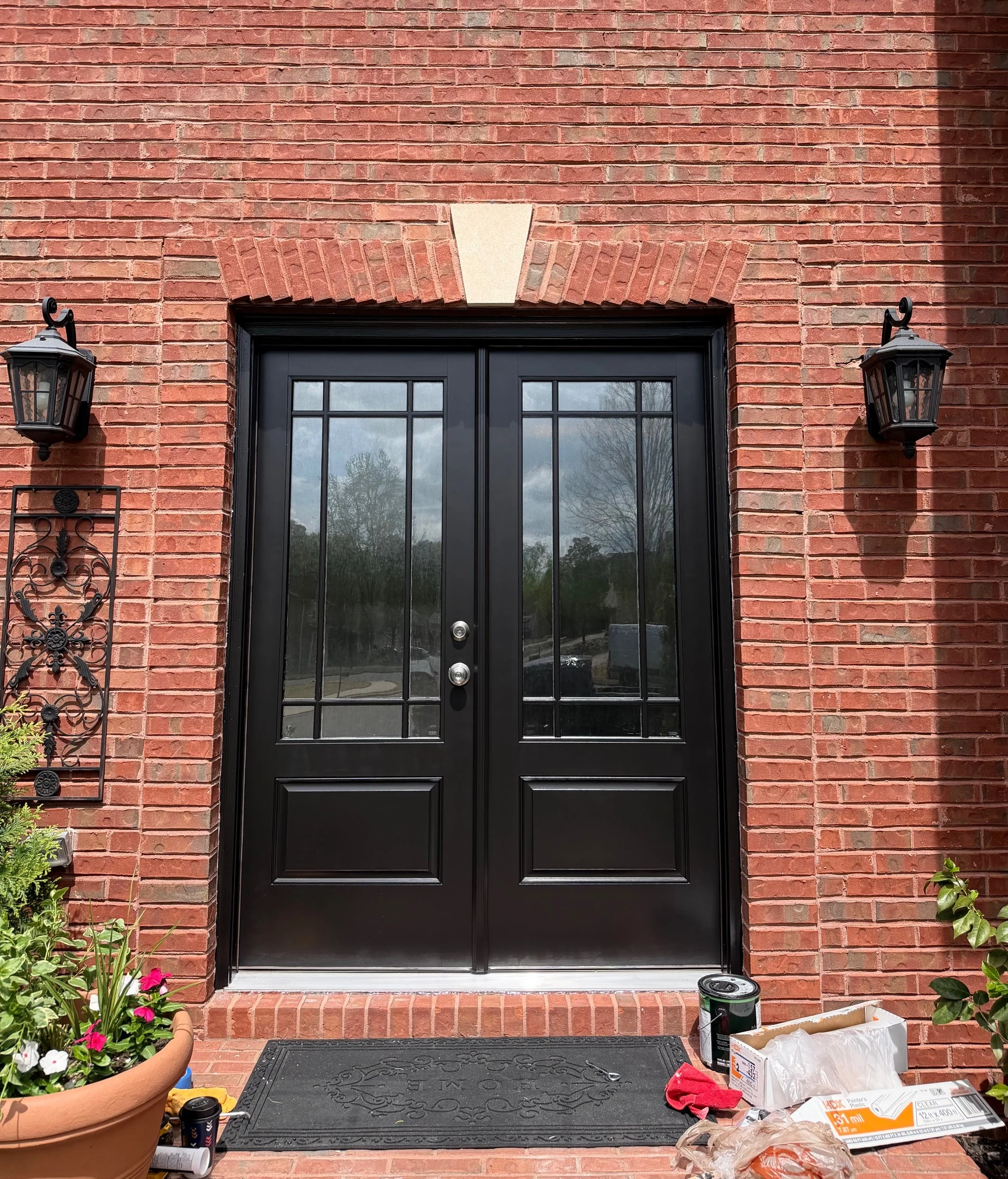Black double door with glass panels in a brick wall, flanked by two black lantern-style wall sconces, a decorative black metal wall hanging on the left, potted plants, and construction supplies near the door.