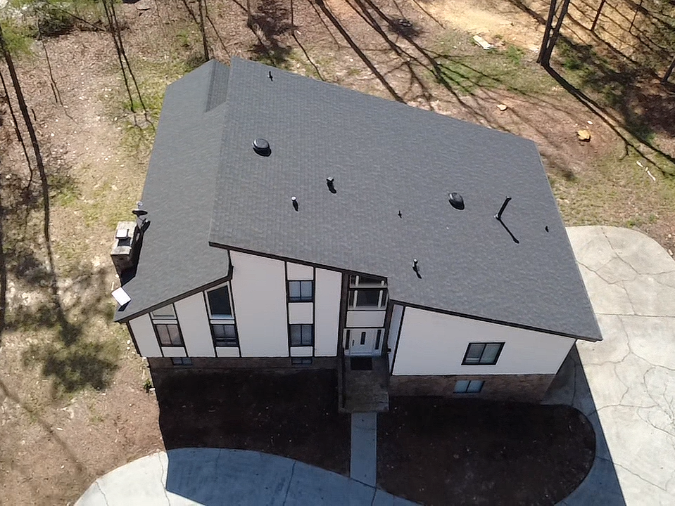 Aerial view of a two-story house with a dark gray roof and white walls, surrounded by a wooded area with some trees and a concrete driveway.