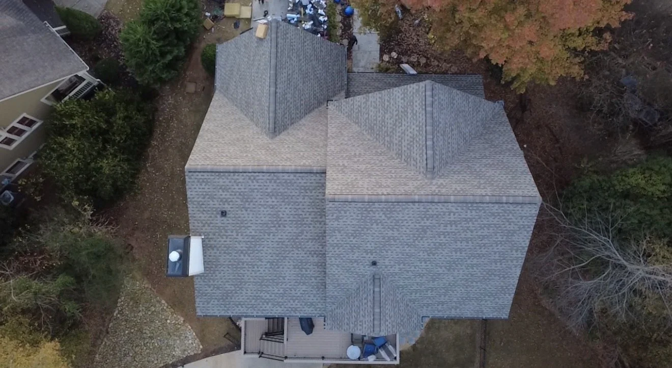 Aerial view of a house with gray shingle roof, surrounded by trees and neighboring houses, with a backyard deck and scattered leaves.