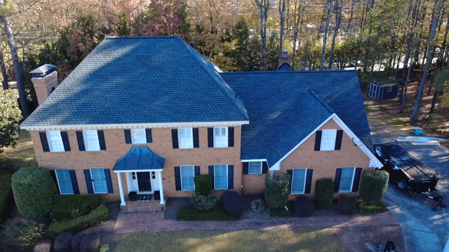 A large brick house with a blue roof, white window shutters, and a front porch with columns, surrounded by well-maintained bushes and trees.