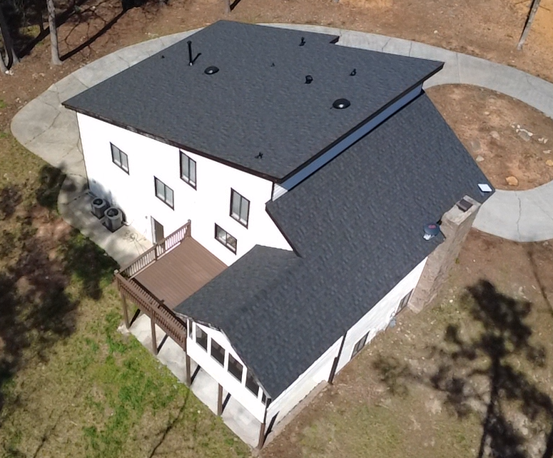 Aerial view of a two-story white house with a dark roof, a small deck, and a paved circular driveway in a yard with grass and bare soil.
