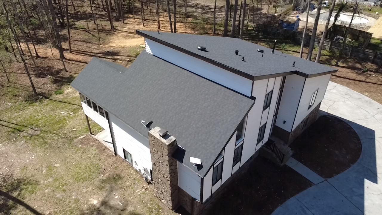 A two-story house with a gray gabled roof, white exterior walls, and large windows, set in a wooded area with a concrete driveway.