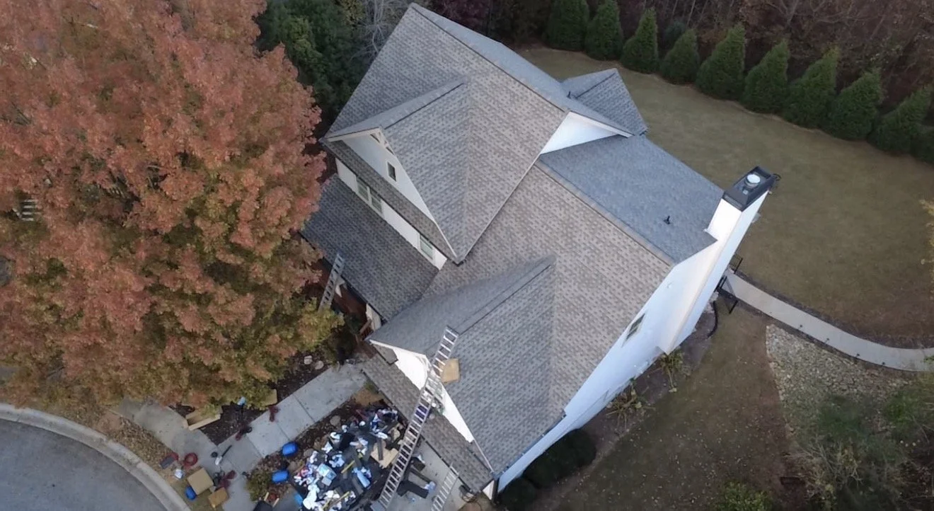 An aerial view of a house with a gray roof, surrounded by trees with autumn-colored leaves, a curved sidewalk, and a yard with a climbing wall and some outdoor furniture.