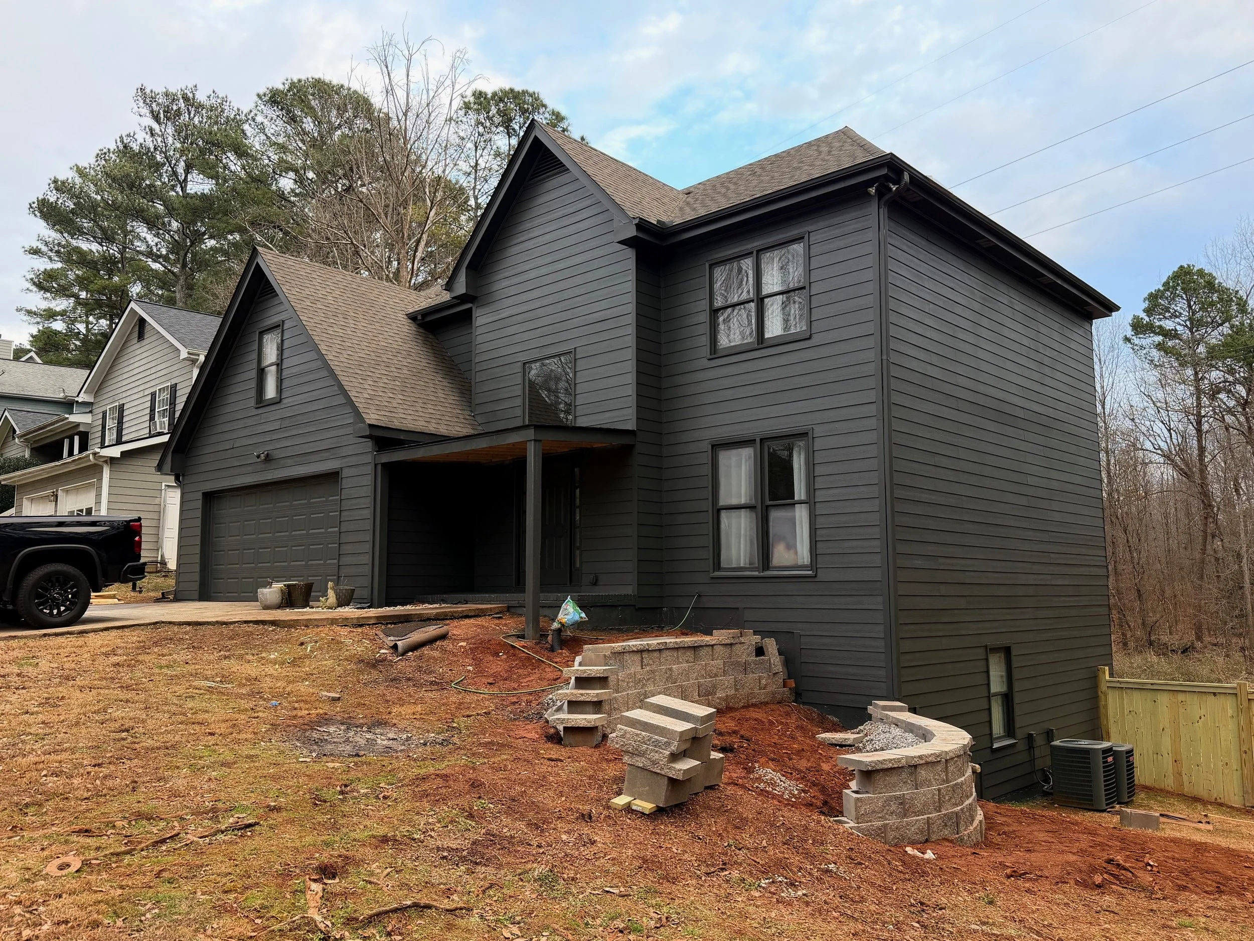 Newly built two-story house with black siding, multiple windows, a garage, and a small porch. Construction is ongoing on the front yard with a small retaining wall and steps, and construction materials are on the ground.