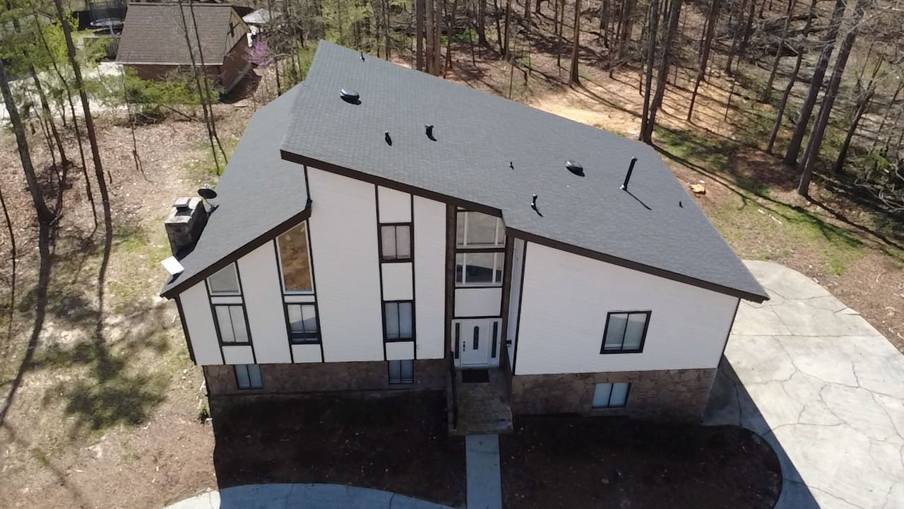A two-story house with a dark gray roof, white siding, and a stone lower facade, surrounded by trees. The house has multiple windows, with some having black trim, and a front door with a small staircase and walkway leading to it. There is a driveway on the right side of the house.